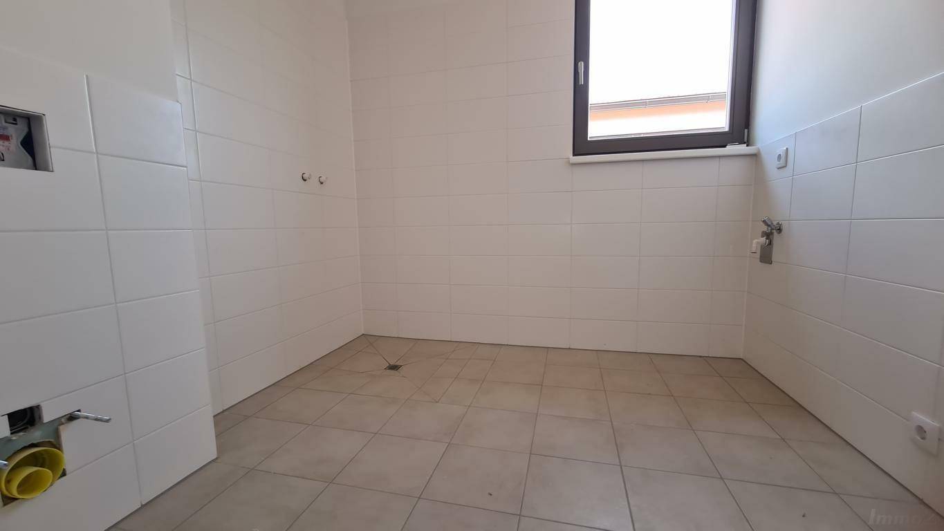 Unmöbliertes Badezimmer mit weißen Fliesen, Fenster und vorbereiteten Anschlüssen für Sanitäranlagen.