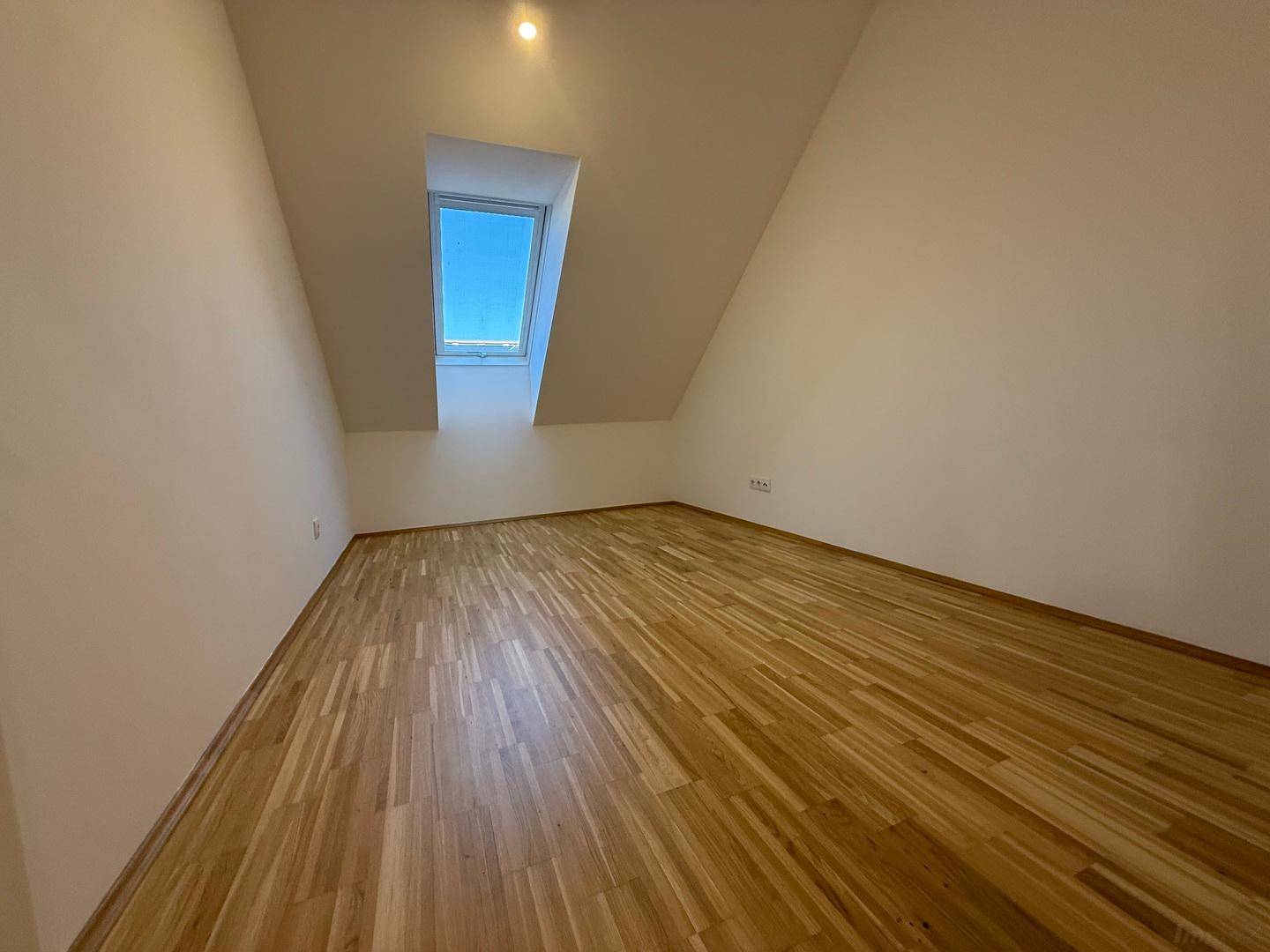 Heller, leerer Raum mit Holzboden, Dachschräge und einem einzelnen Dachfenster.