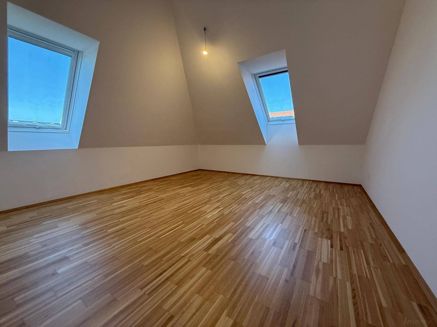 Leeres Schlafzimmer mit Holzboden, Dachschrägen und zwei hellen Dachfenstern.