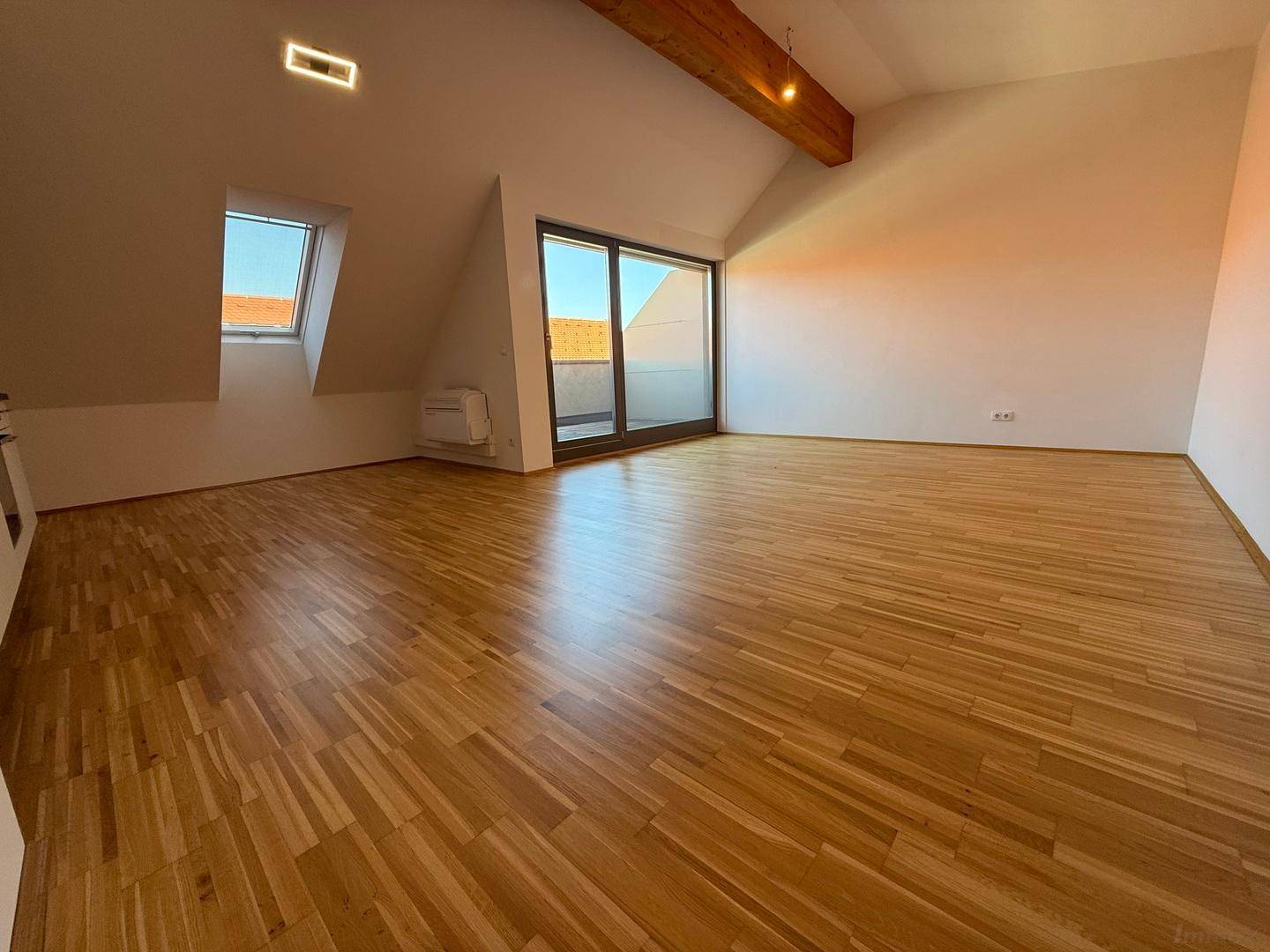 Geräumiger, leerer Wohnbereich mit hellem Parkettboden, Dachschrägen und Zugang zur Terrasse.