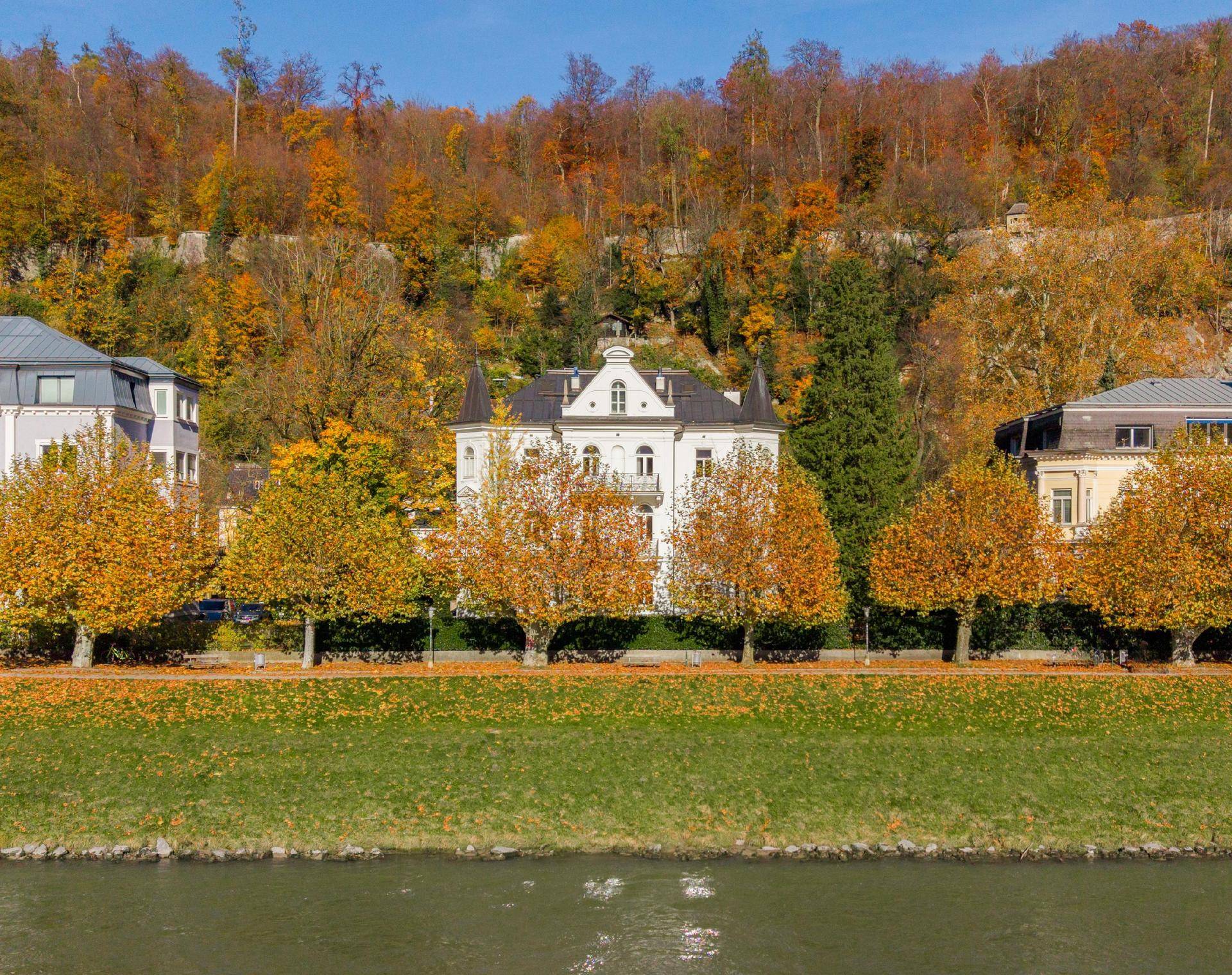 Elegante weiße Villa am Flussufer, umgeben von herbstlichen Bäumen und einem bewaldeten Hügel im Hintergrund.