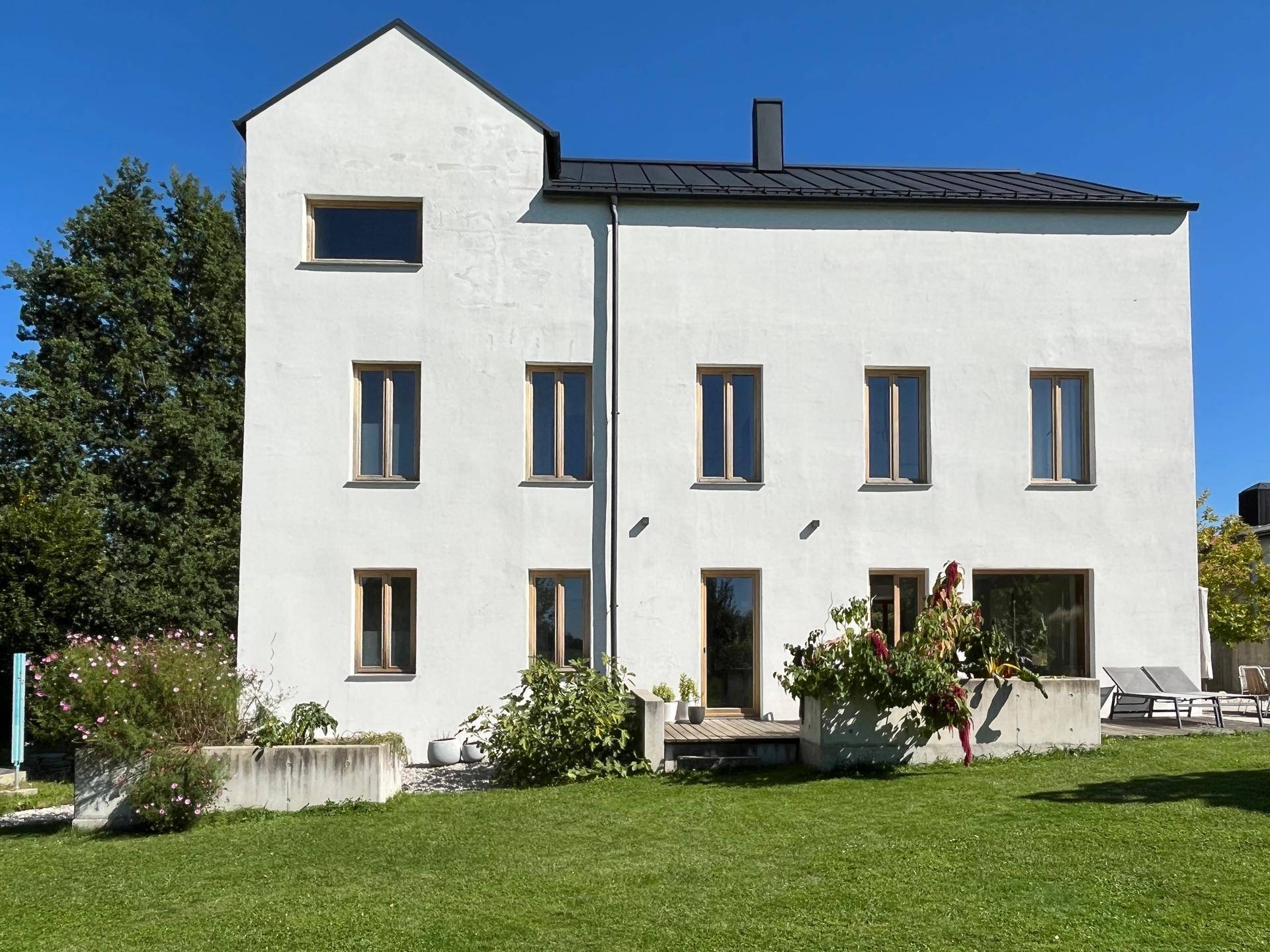 Rückansicht des modernen weißen Gebäudes mit Rasenfläche und kleiner Terrasse unter blauem Himmel.
