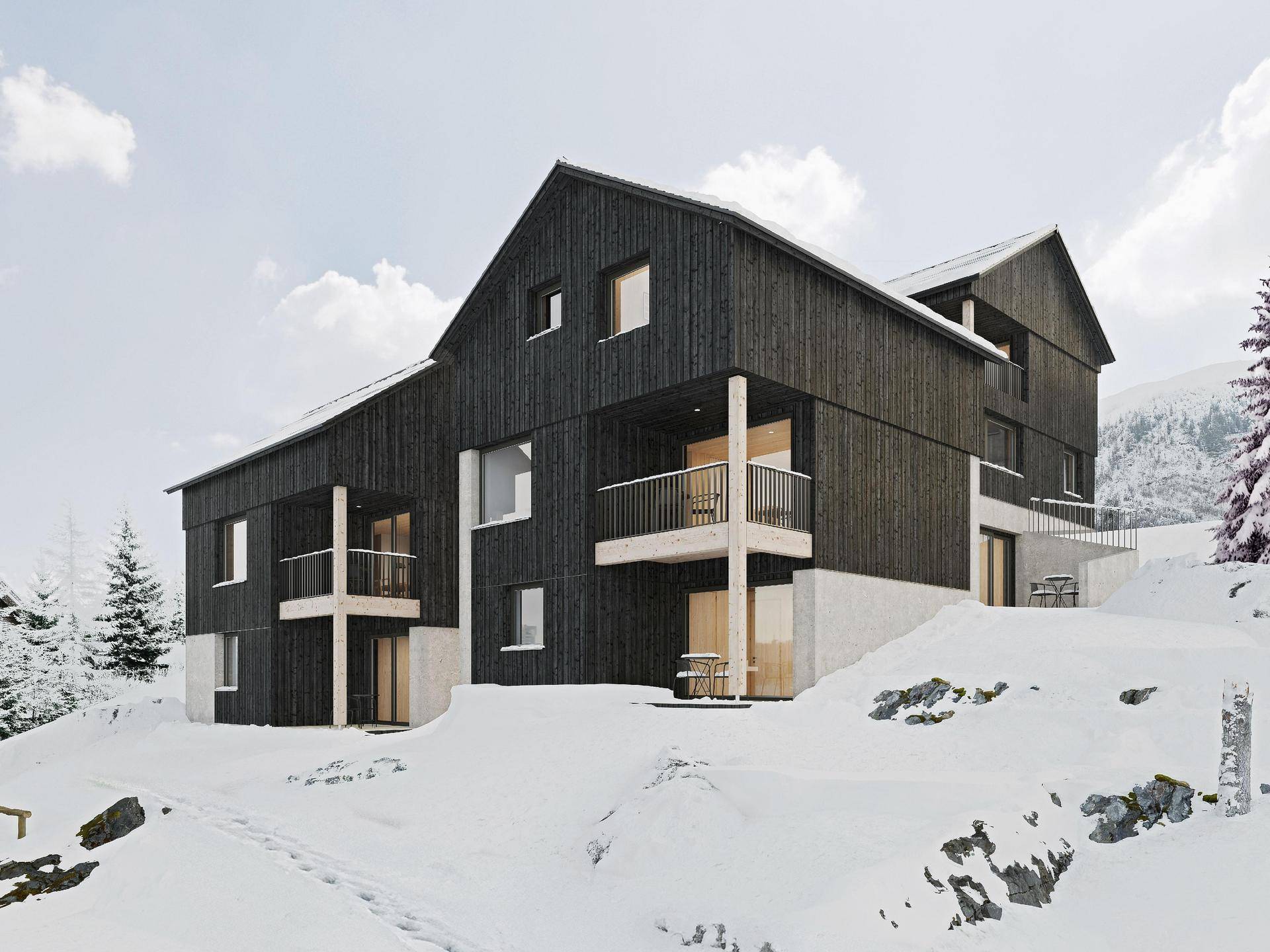 Modernes Gebäude mit dunkler Holzfassade und Balkonen, eingebettet in eine verschneite Berglandschaft.
