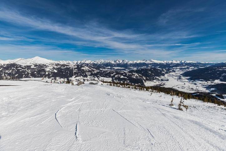 Weitläufige, verschneite Berglandschaft mit Skipisten und einem weiten Talblick unter blauem Himmel.