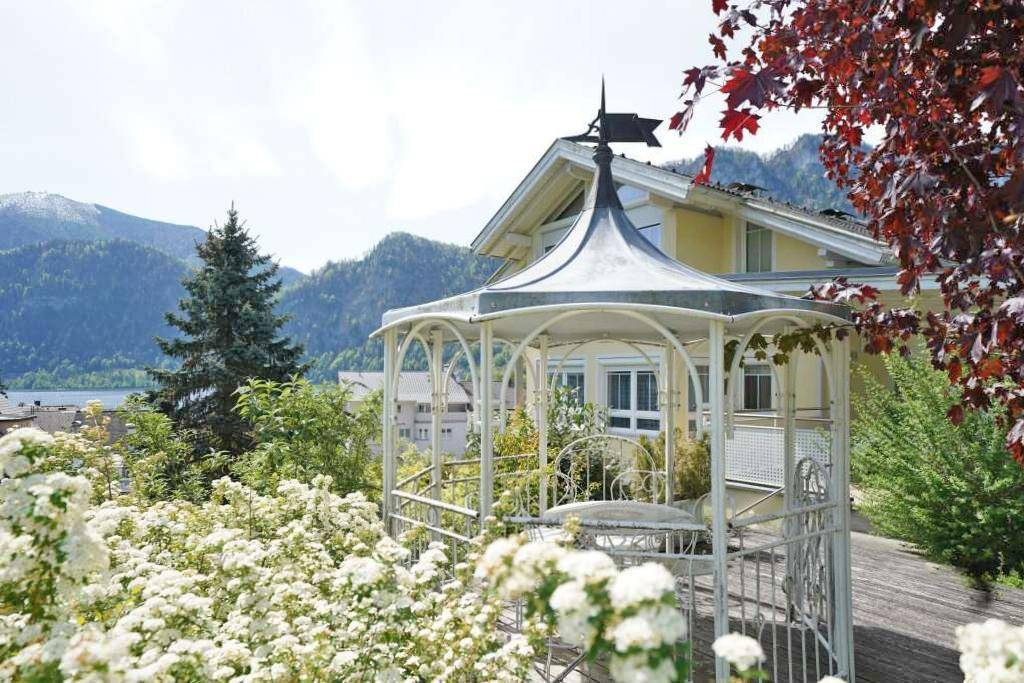 Romantischer Pavillon im Garten mit weissen Blüten und malerischem Blick auf den See und die Berge.