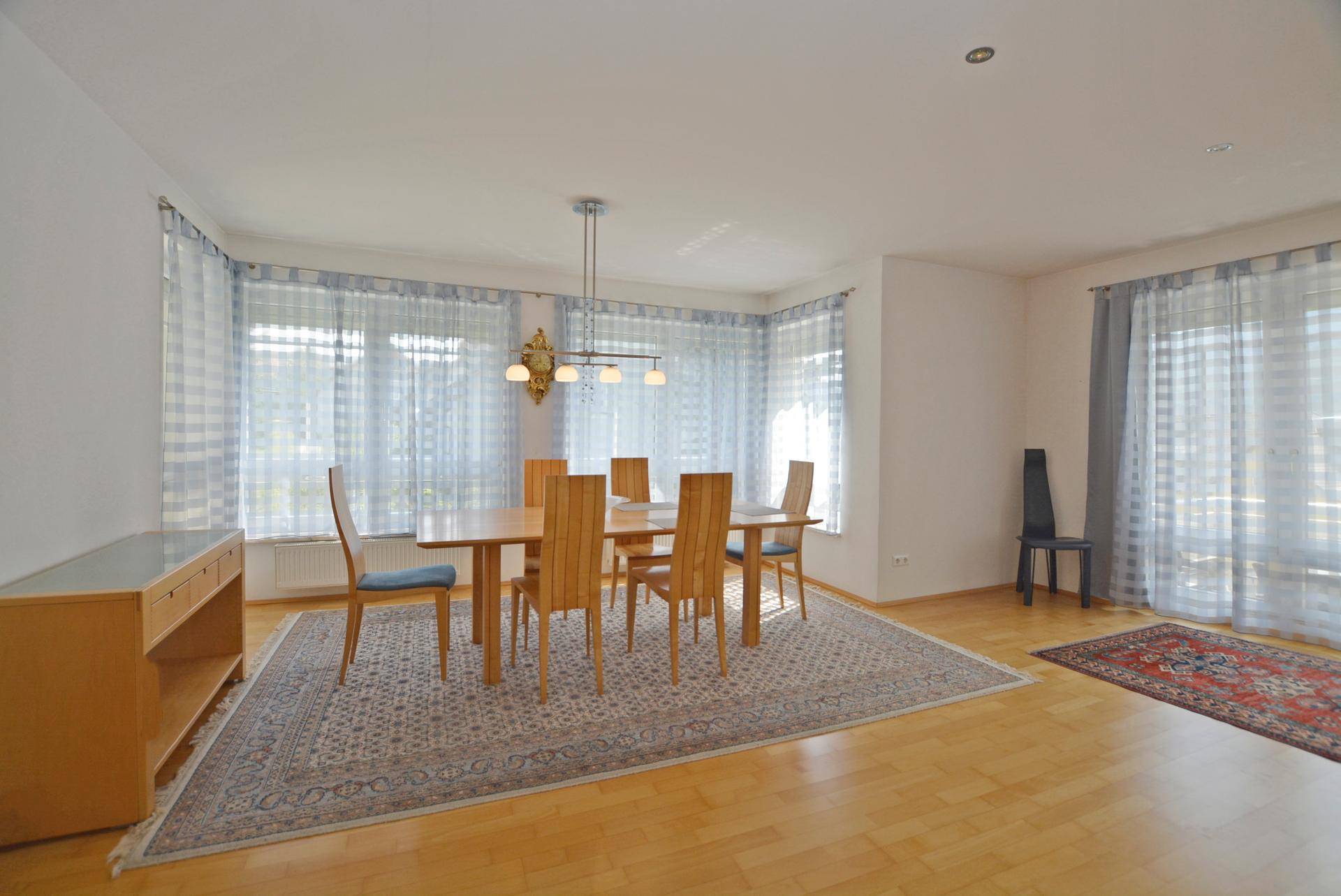 Heller Essbereich mit Holztisch, passenden Stühlen und großen Fenstern mit hellblauen Vorhängen.