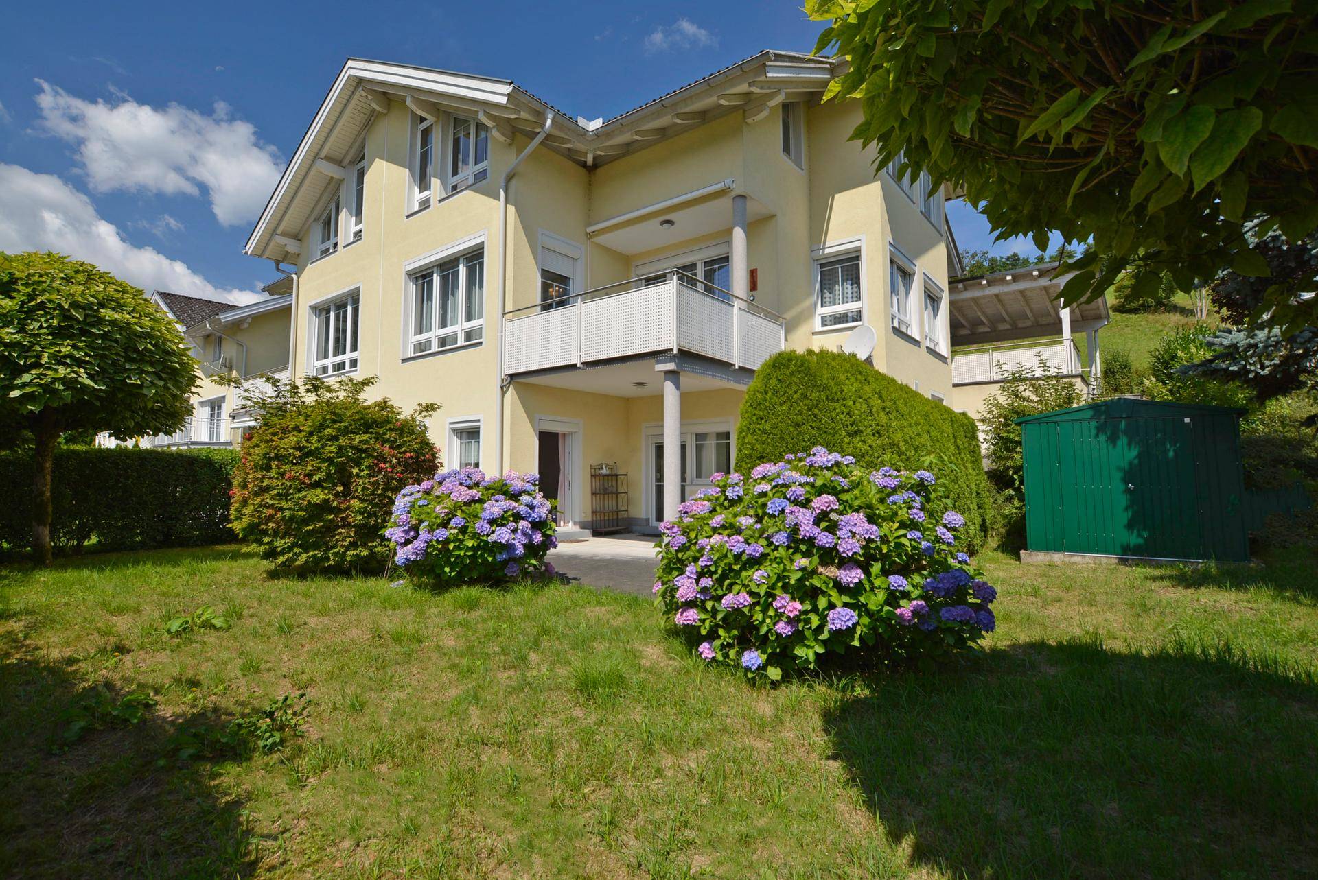 Vorderansicht des Hauses mit gepflegtem Rasen, blühenden Hortensien und einer kleinen Terrasse.