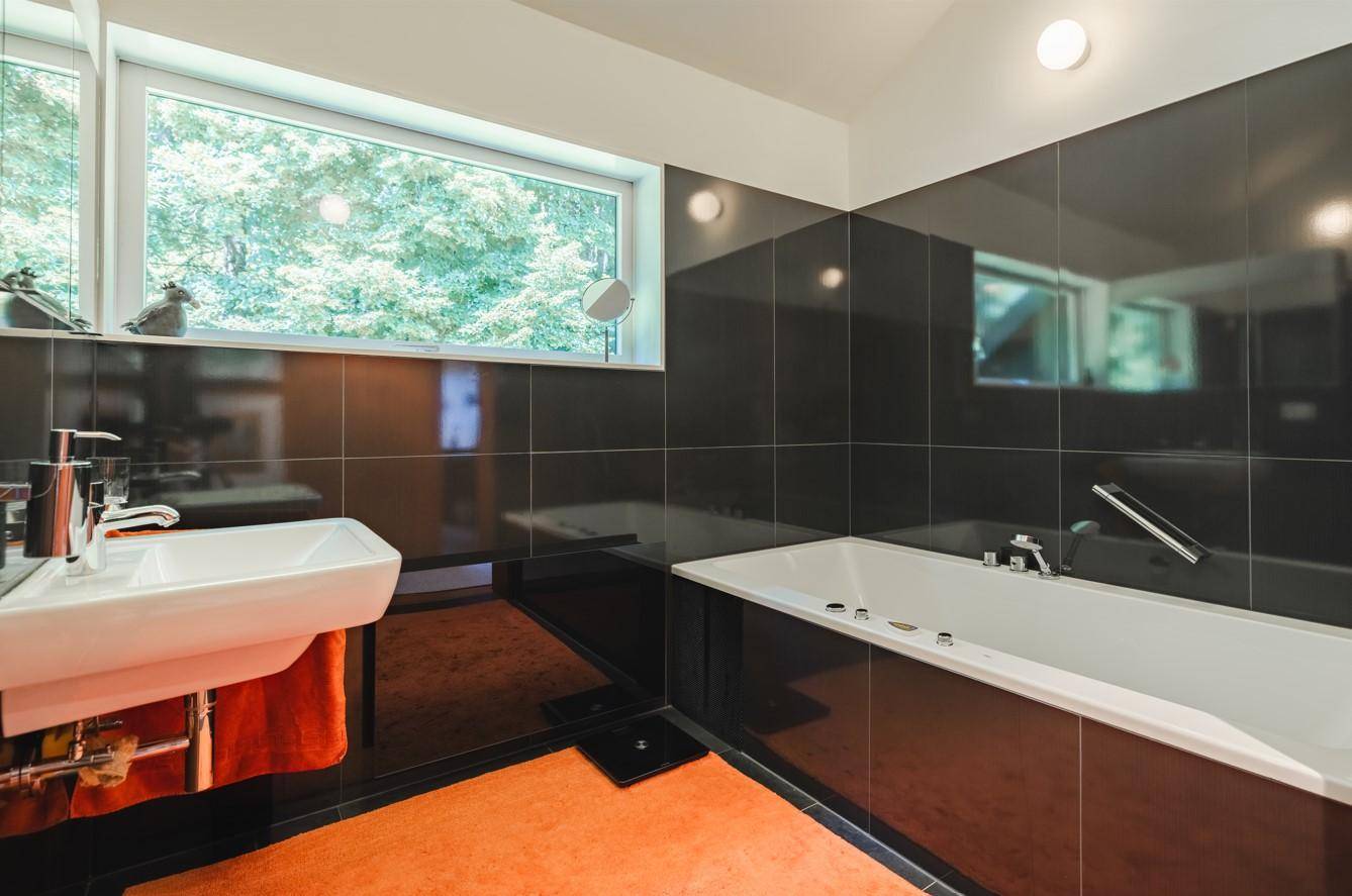 Modernes Badezimmer mit schwarzen Hochglanzfliesen, Badewanne und Fenster mit Blick ins Grüne.
