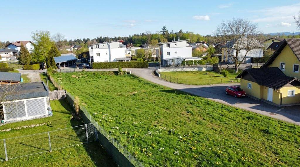 Grünes Baugrundstück mit angrenzender Straße und verschiedenen Wohnhäusern unter blauem Himmel.