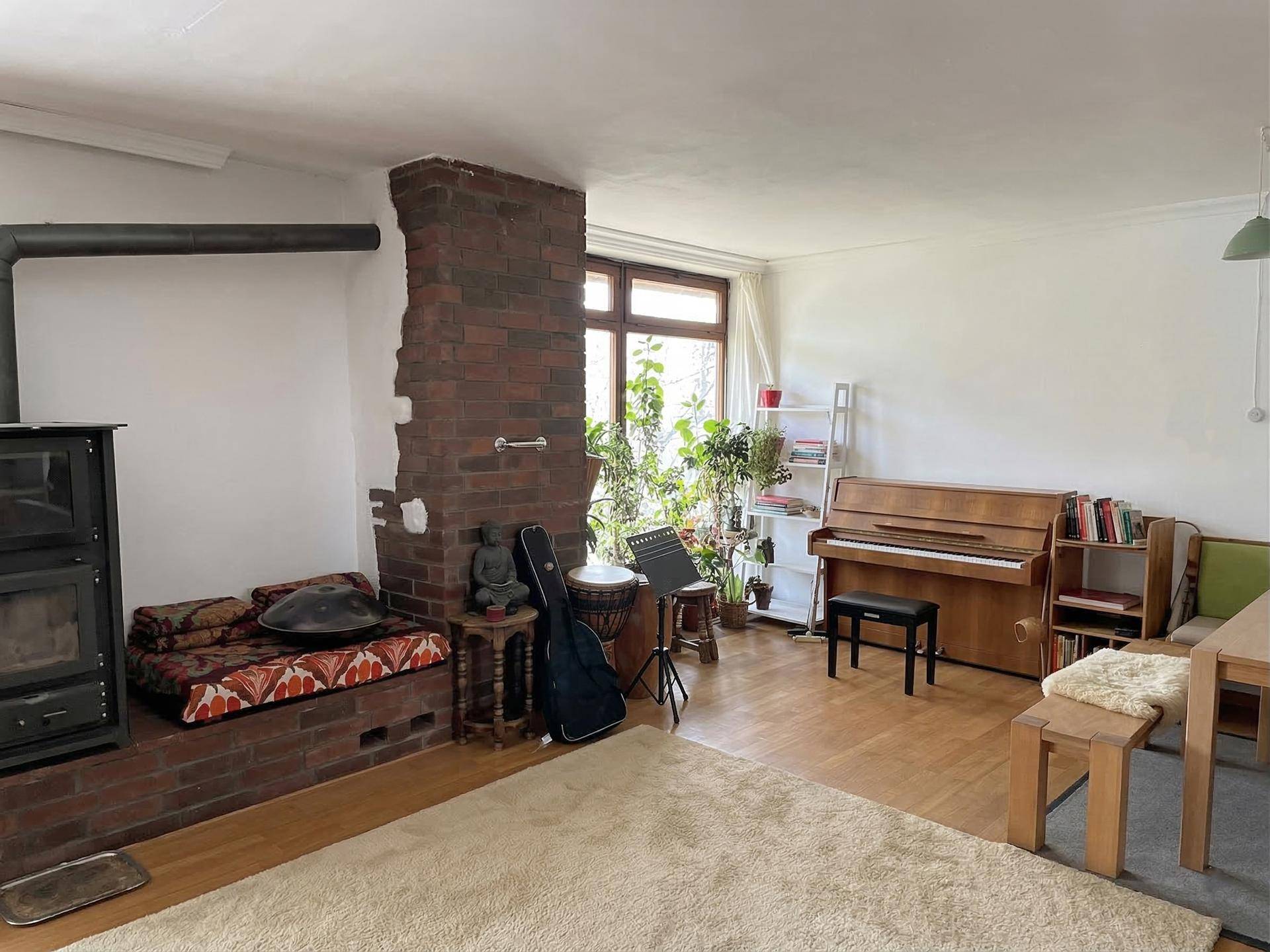 Heller Wohnbereich mit Kaminofen, Ziegelwandelement und Klavier, umgeben von Pflanzen.