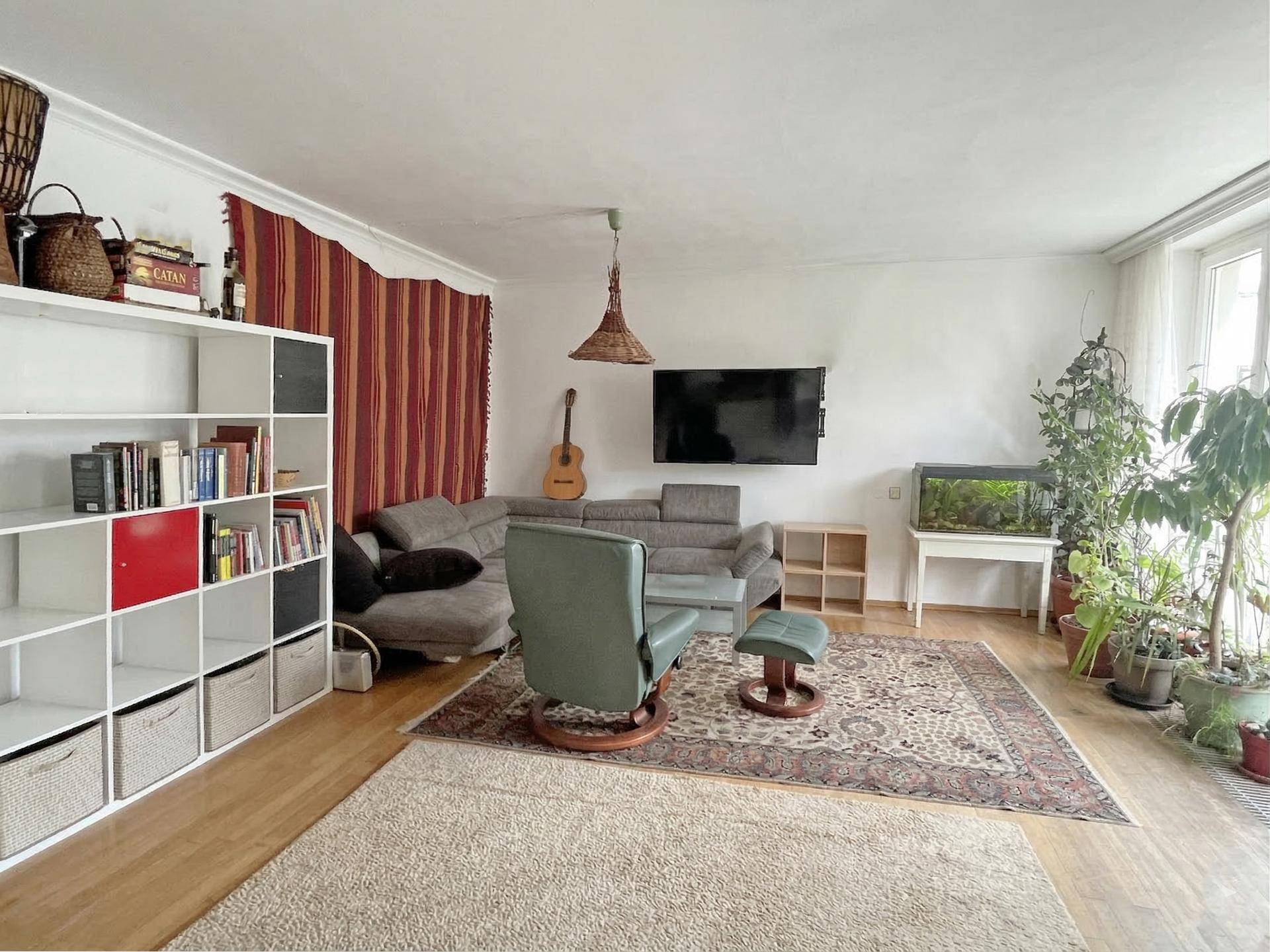 Gemütliches Wohnzimmer mit Sofa, Sessel, Bücherregal und vielen Pflanzen, hell und einladend.