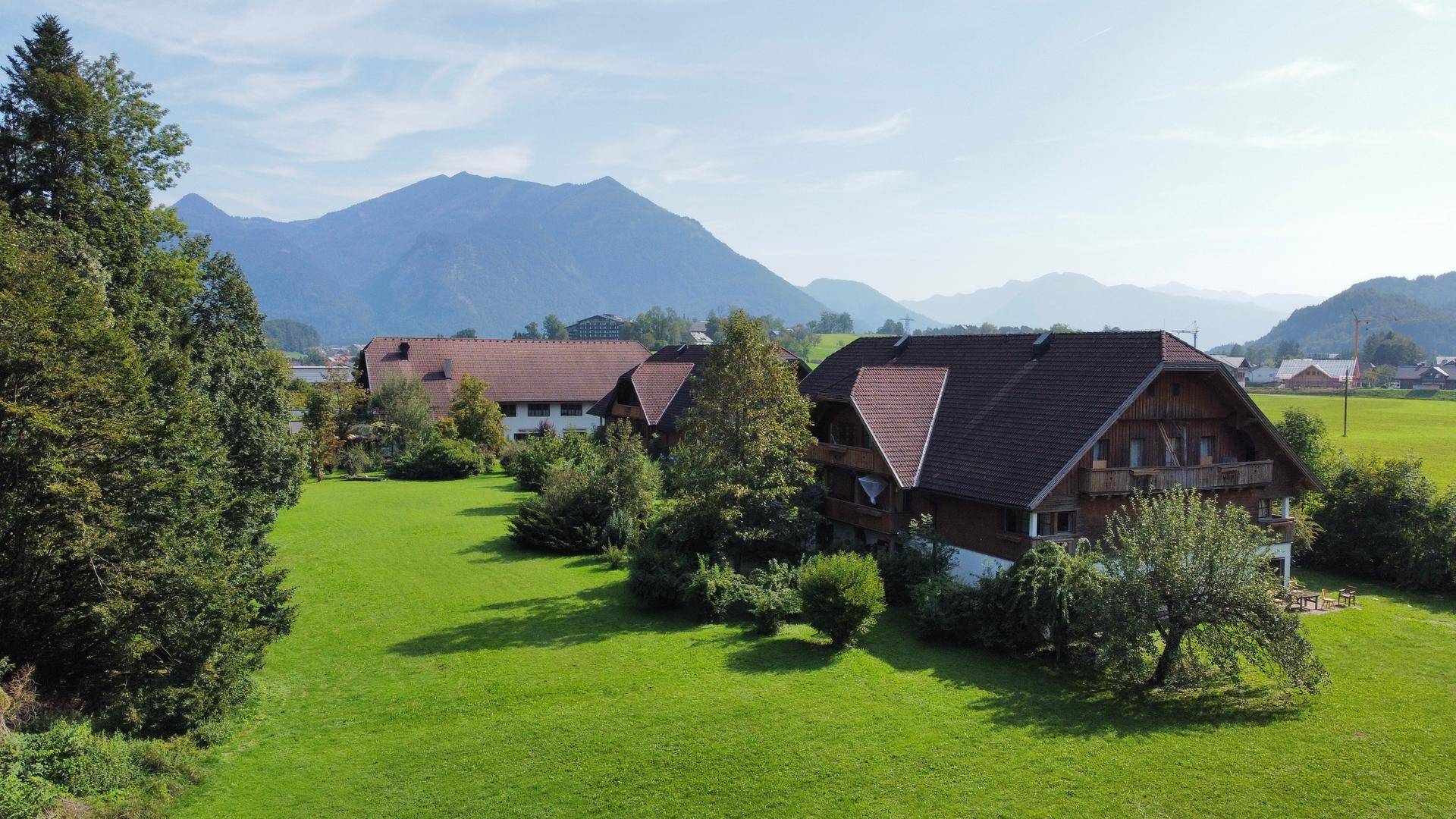 Traditionelle Häuser mit braunen Dächern, umgeben von weitläufigen grünen Wiesen und Bäumen vor Bergkulisse.