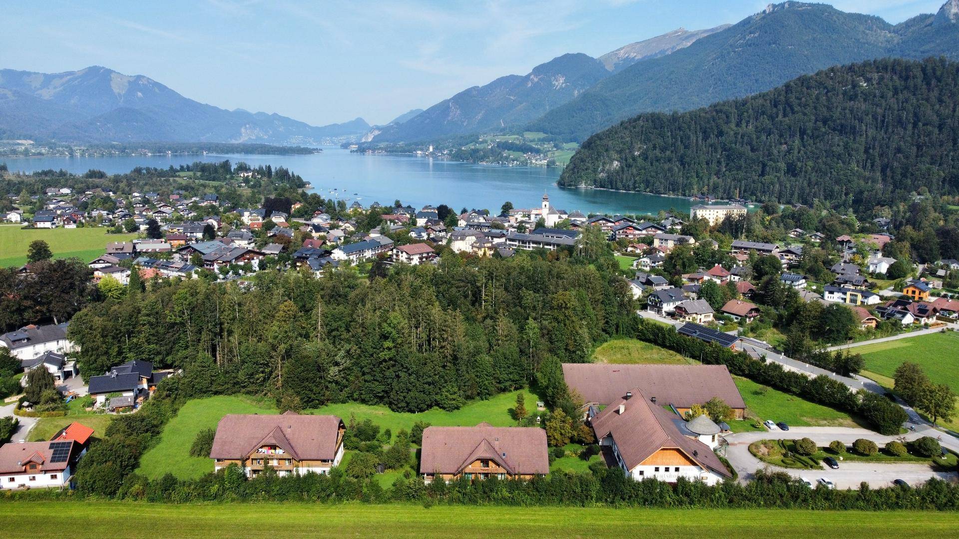 Luftaufnahme einer Siedlung am Seeufer mit traditionellen Häusern, eingebettet in grüne Felder und bewaldete Hügel.