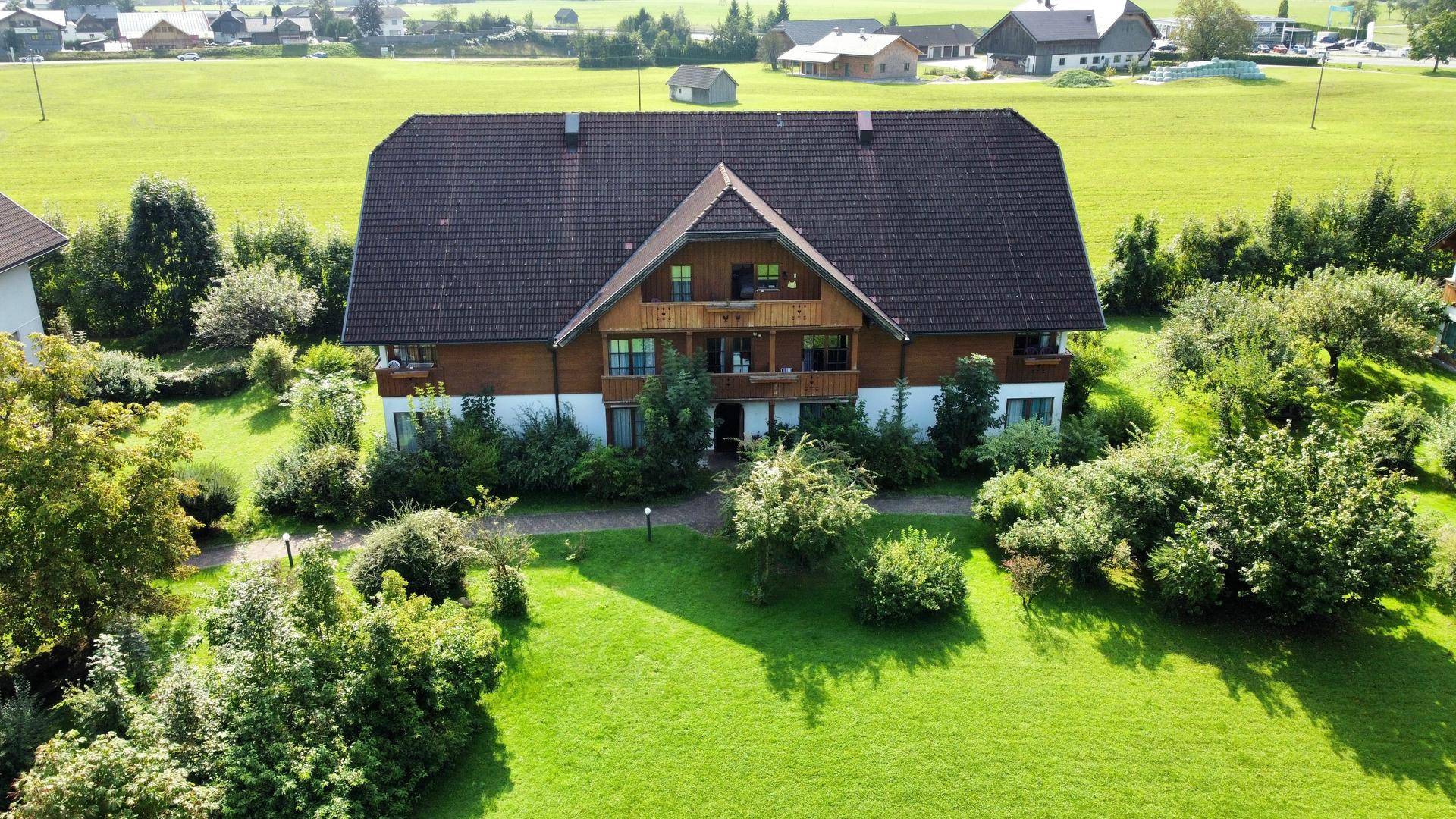 Großes Haus mit Holzfassade und braunem Dach, umgeben von einem gepflegten grünen Garten mit Bäumen und Sträuchern.
