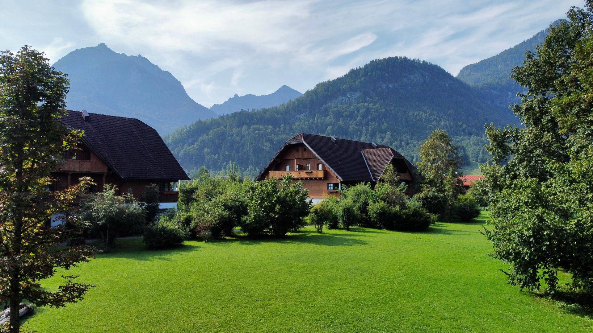 Idyllische Häuser in einer weiten grünen Wiese, umrahmt von üppigen Bäumen und hohen Bergen im Hintergrund.