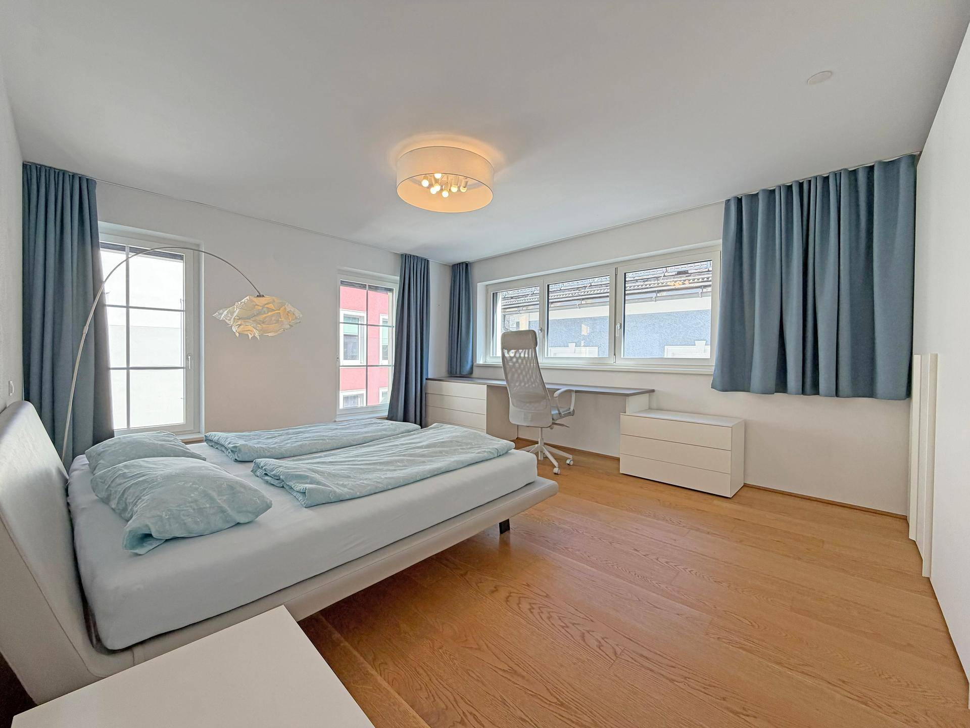 Geräumiges Schlafzimmer mit Holzboden, Schreibtisch und großen Fenstern mit blauen Vorhängen.
