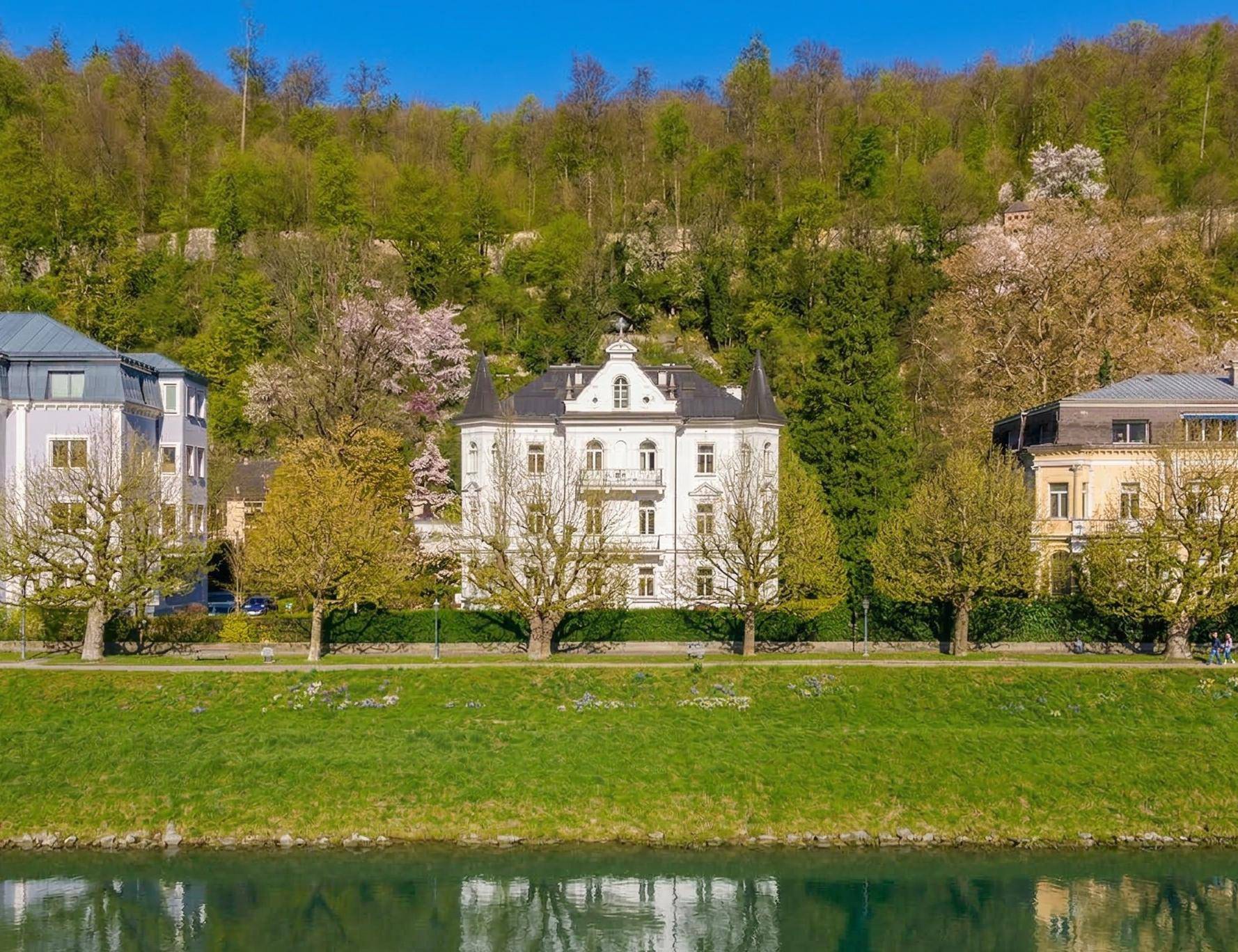 Panoramablick auf die historische Villa am Flussufer mit bewaldetem Hügel im Hintergrund.