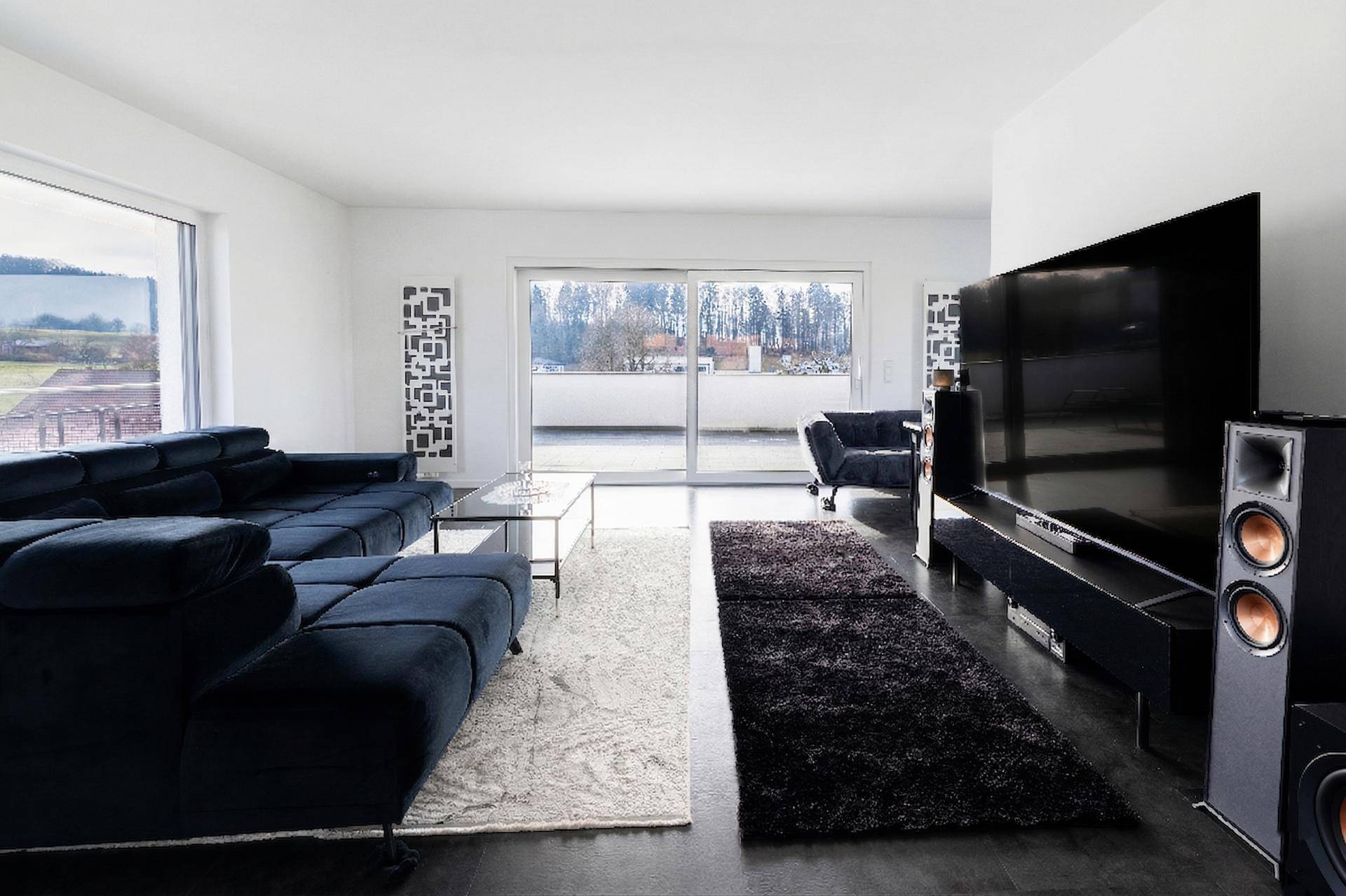 Großzügiges Wohnzimmer mit dunklem Sofa, großem Fernseher und Zugang zur Terrasse durch Schiebetüren.
