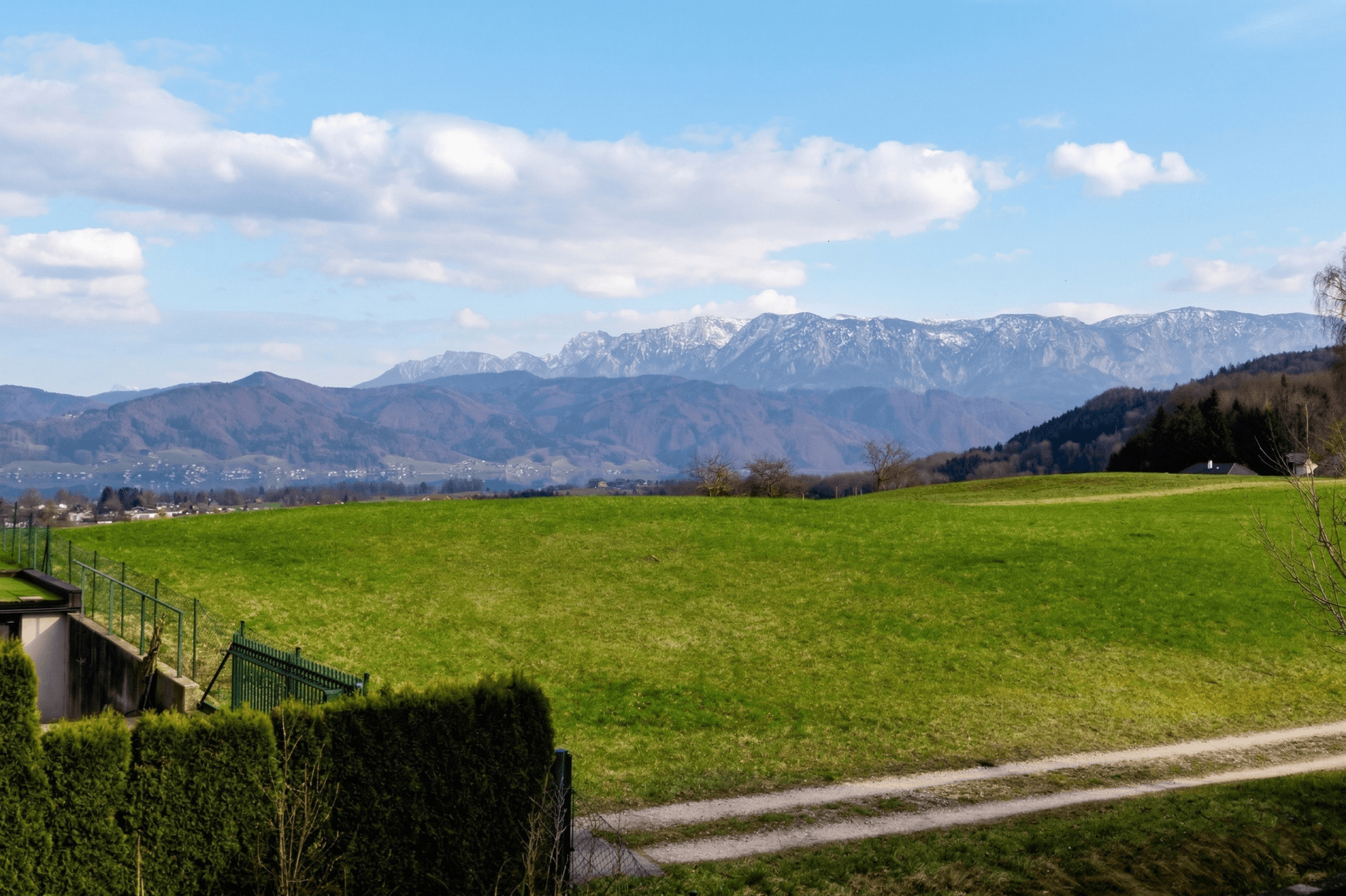 Weitläufiger Blick über grüne Hügel, einen See und majestätische Berge am Horizont.