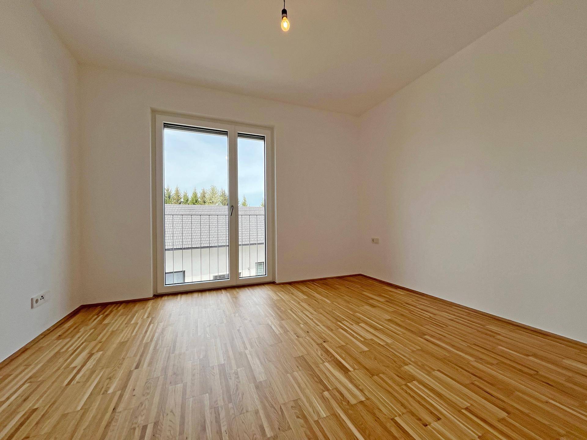 Helles Schlafzimmer mit Parkettboden und großem Fenster, das viel Tageslicht hereinlässt.