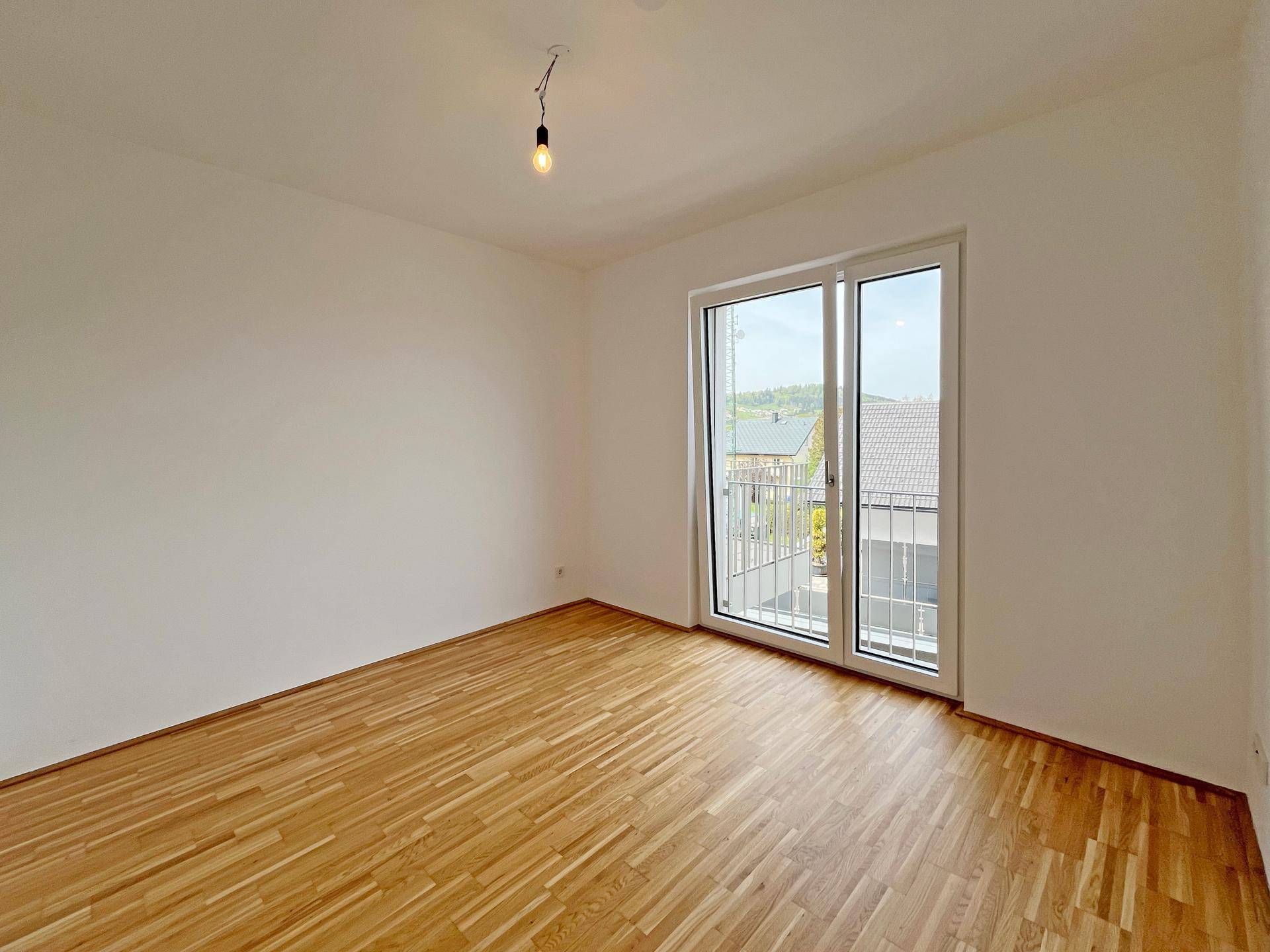 Weiteres helles Schlafzimmer mit Parkettboden und einem Fenster mit Blick ins Grüne.