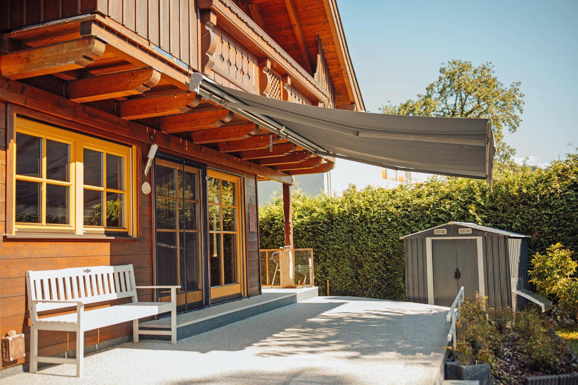 Gemütliche Terrasse am Holzhaus mit weißer Bank, ausfahrbarer Markise und einem kleinen Gartenschuppen.