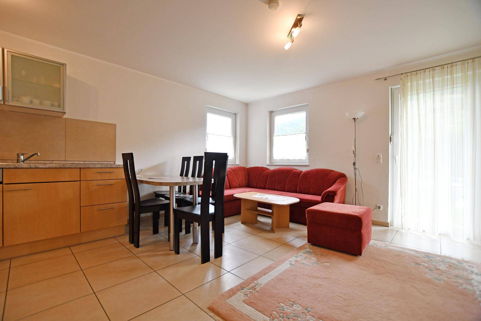 Gemütlicher Wohn-Essbereich mit integrierter Küchenzeile, rotem Sofa und Zugang zur Terrasse.