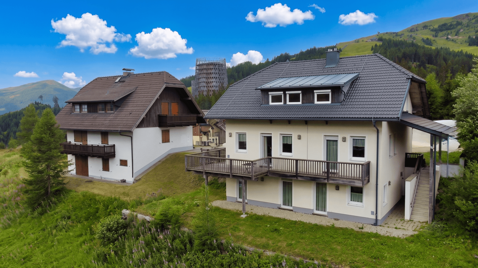 Zwei Häuser auf einem grünen Hügel mit moderner Architektur im Hintergrund und Bergblick.