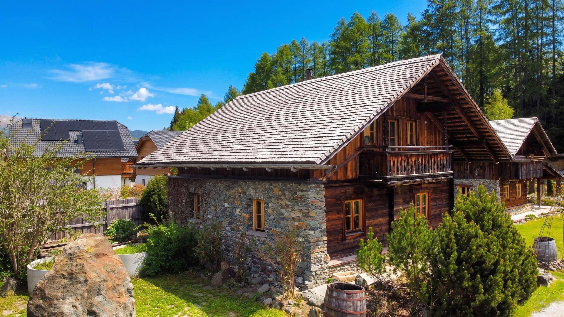 Rustikales Holzhaus mit Steinfundament und Schindeldach, umgeben von grüner Natur und Bergen.