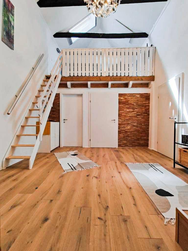Heller offener Raum mit Holztreppe zur Galerie, Holzboden und dekorativer Wandverkleidung.