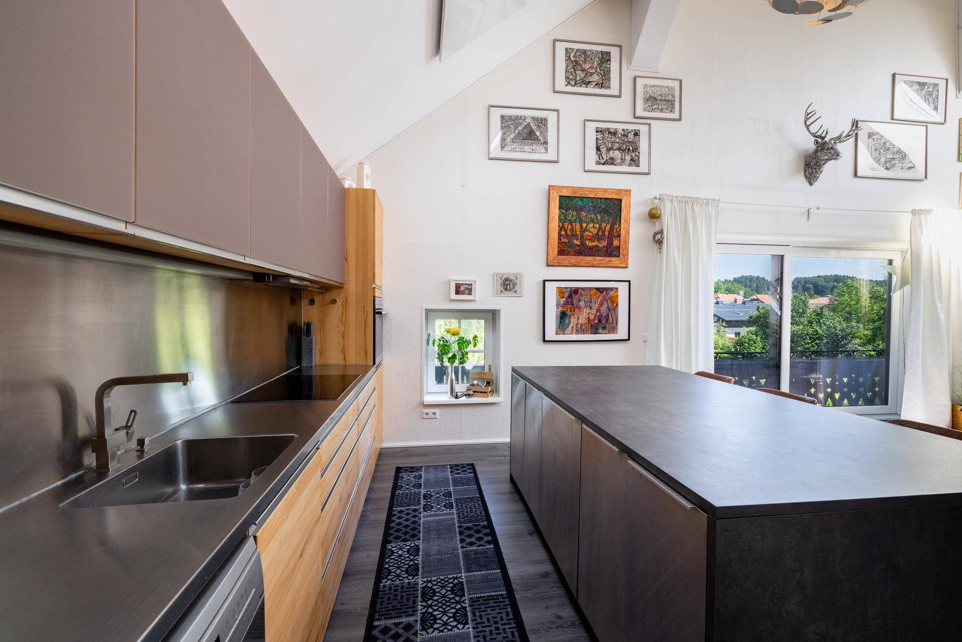 Moderne Küche mit Edelstahl-Arbeitsplatten, Holzelementen und großem Fenster mit grüner Aussicht.