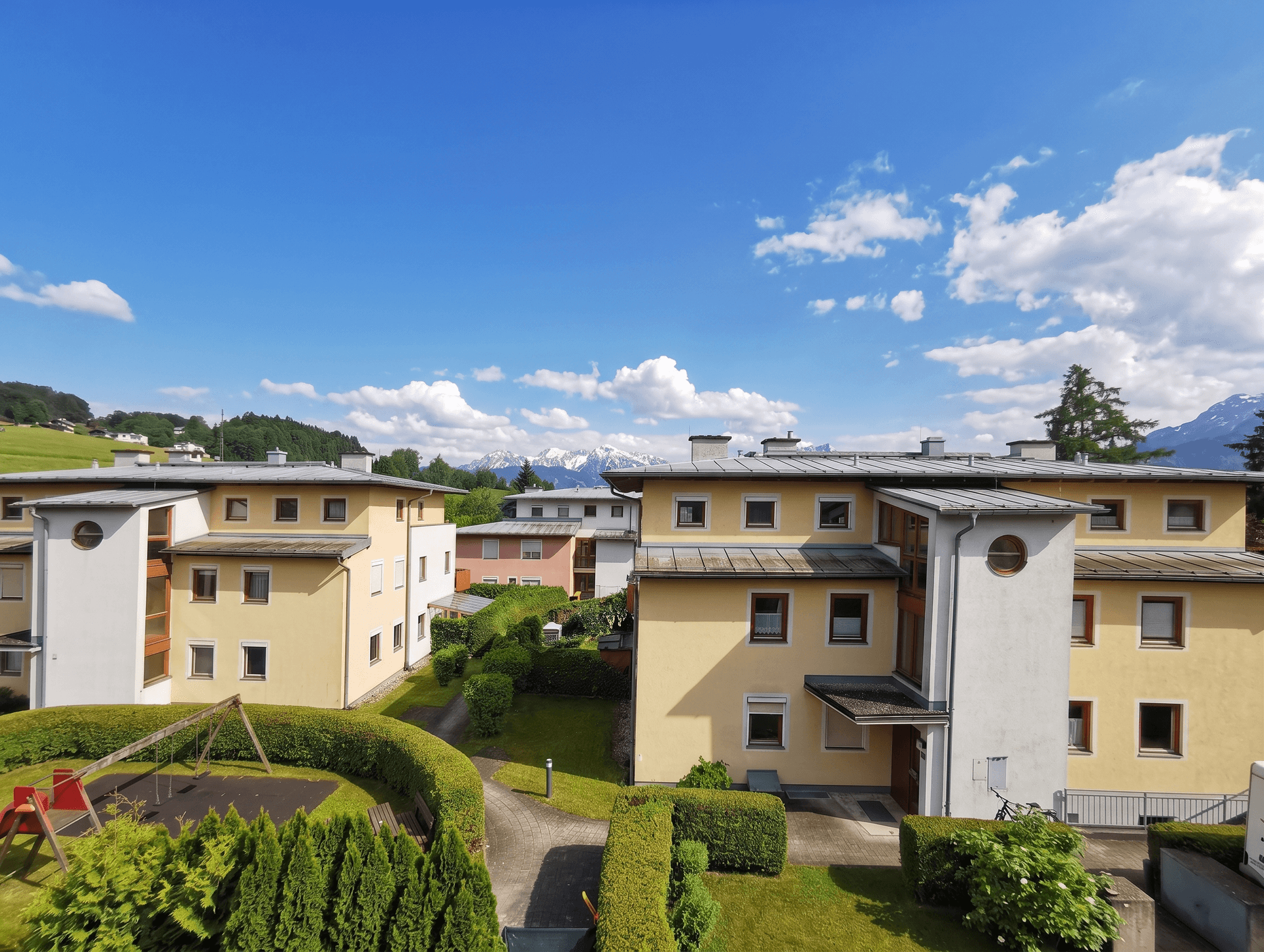 Wohnanlage mit gepflegten Grünflächen, Spielplatz und Bergblick unter blauem Himmel.