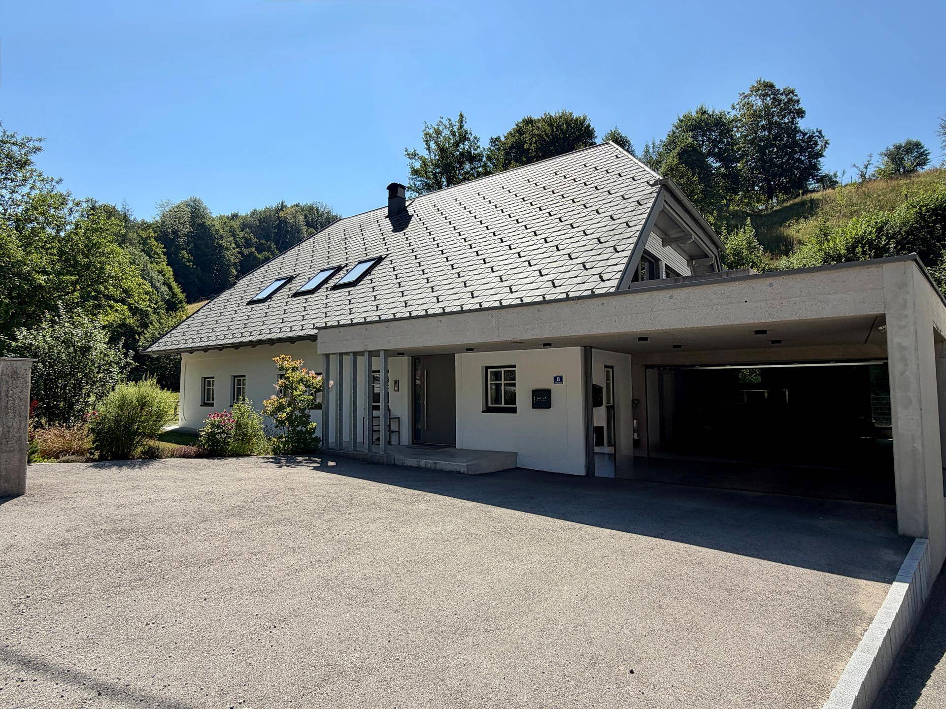 Modernes Haus mit Satteldach und integriertem Carport, umgeben von Bäumen unter blauem Himmel.