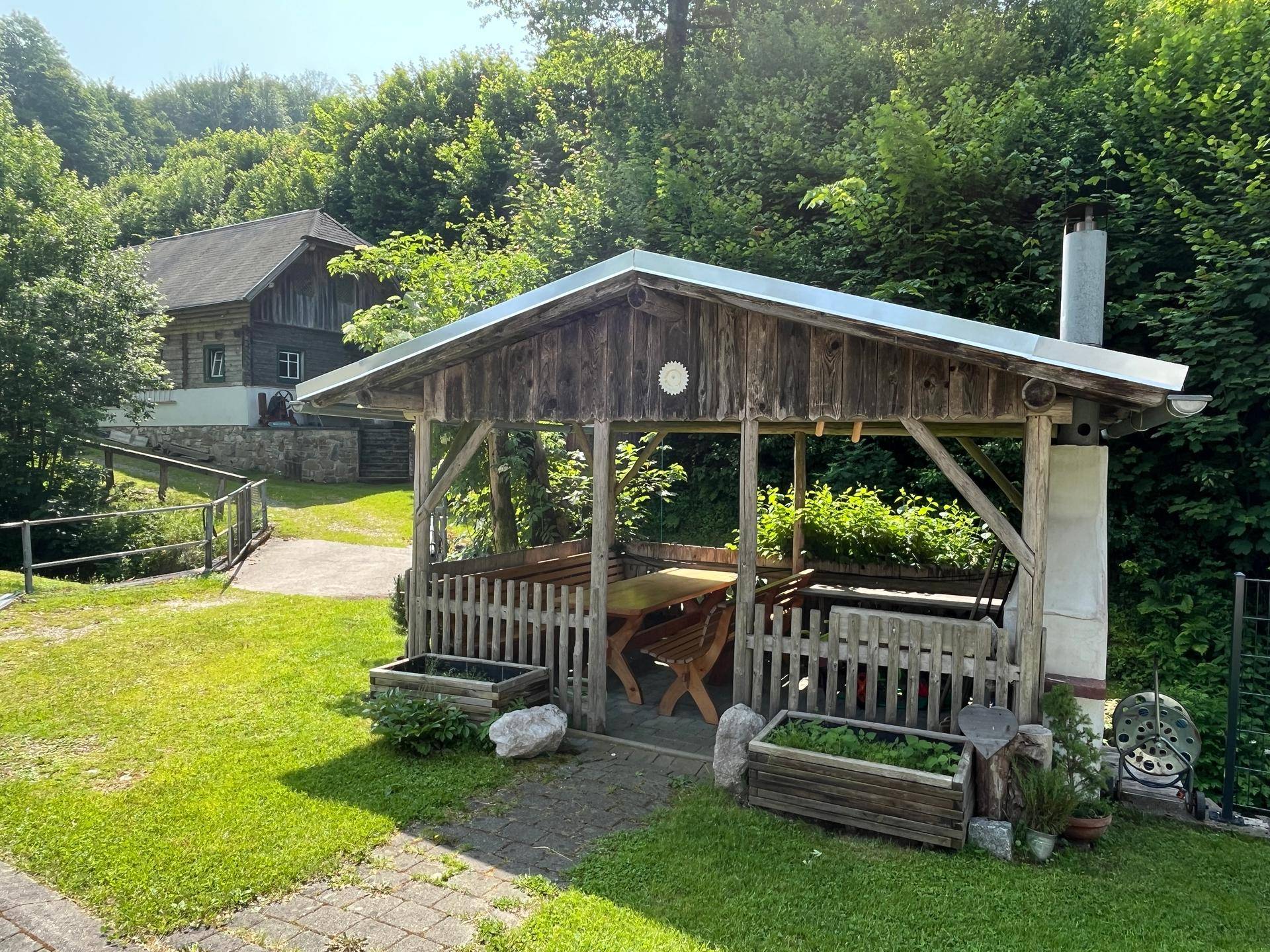 Gemütlicher Holzpavillon im Garten mit Sitzgelegenheiten und Grillplatz, umgeben von Grün.