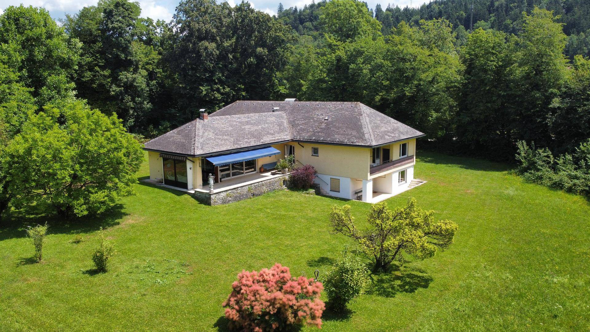 Luftaufnahme eines L-förmigen Hauses mit großem Garten und umliegenden Wäldern.
