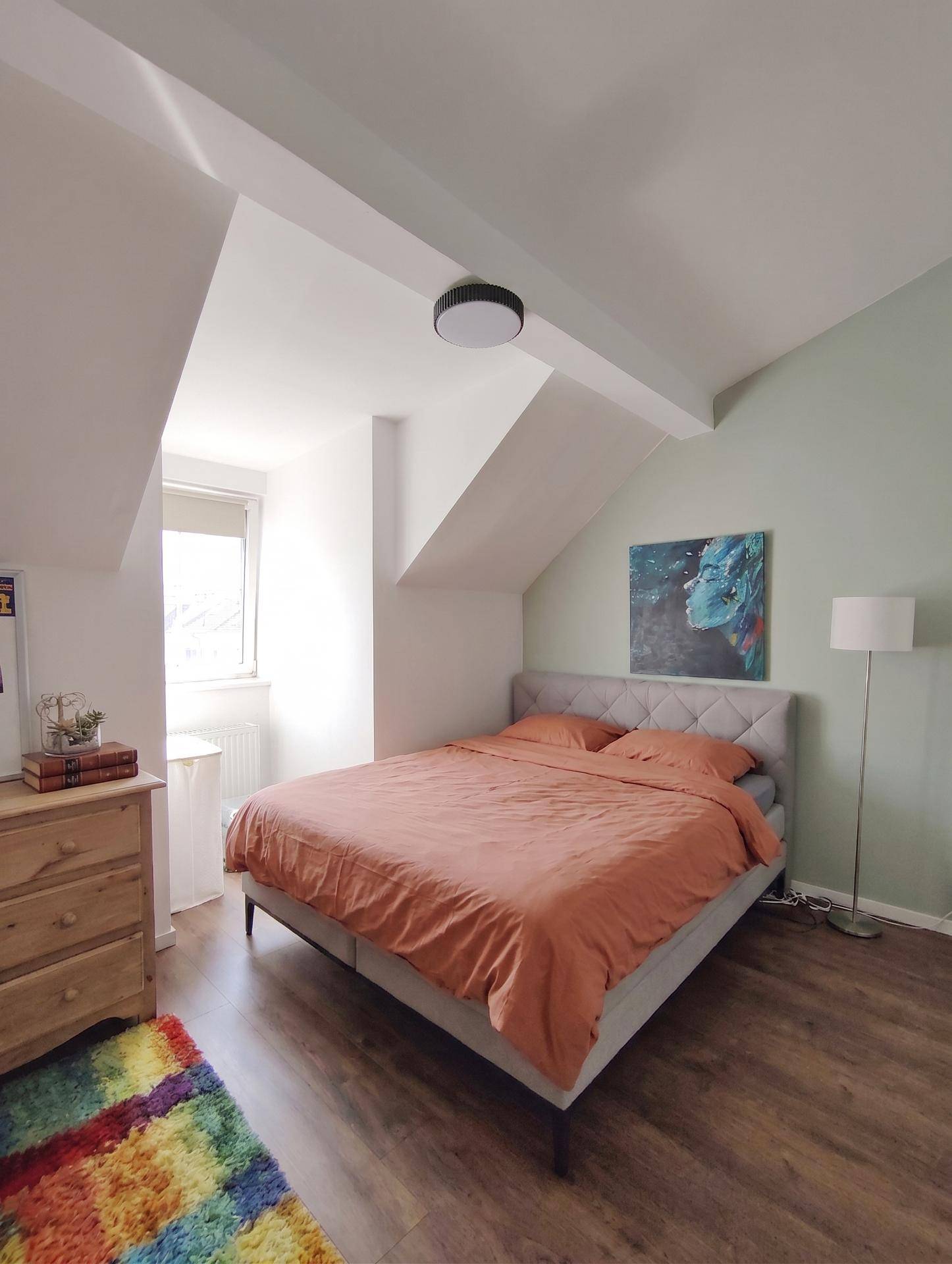 Schlafzimmer im Dachgeschoss mit gemütlichem Bett, schrägen Wänden und einem bunten Teppich.