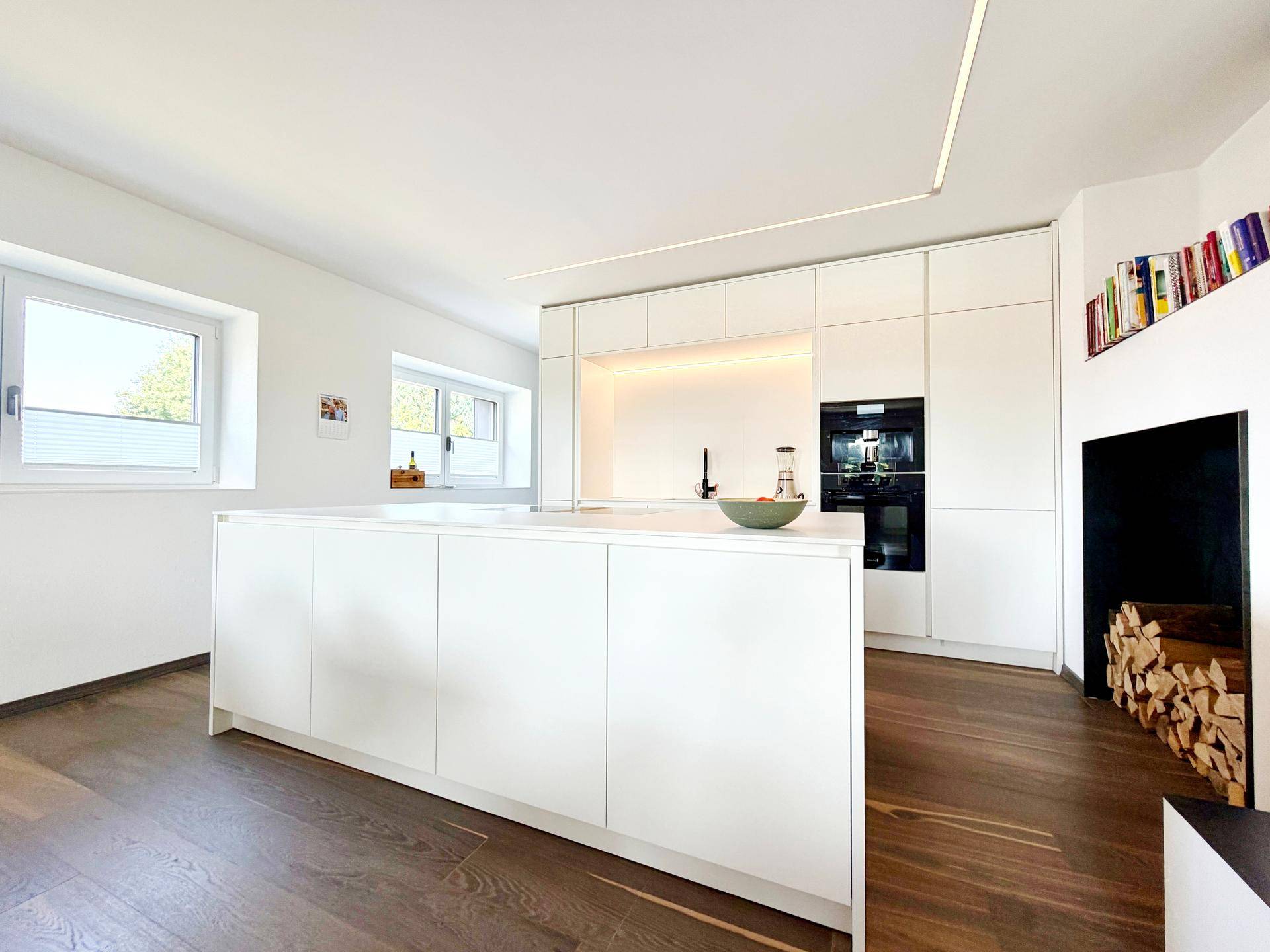 Moderne weiße Küche mit Kochinsel, integrierten Geräten und Holzlager neben dem Kamin.