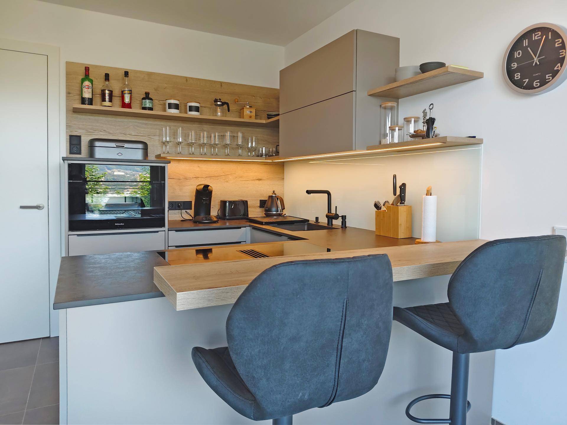 Moderne Küche mit grauen Schränken, Holzakzenten, integriertem Backofen und einer praktischen Frühstücksbar mit Hockern.