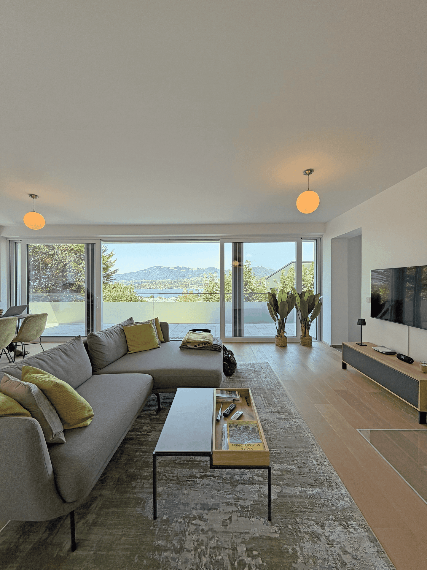 Geräumiges Wohnzimmer mit großem Sofa, Fernseher und bodentiefen Fenstern mit Panoramablick auf See und Berge.