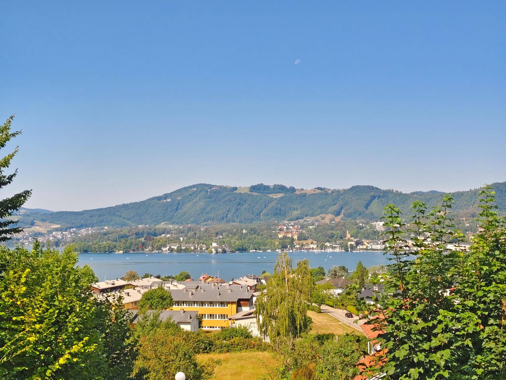 Malerischer Panoramablick auf einen See, umgeben von grünen Bergen und Siedlungen unter blauem Himmel.