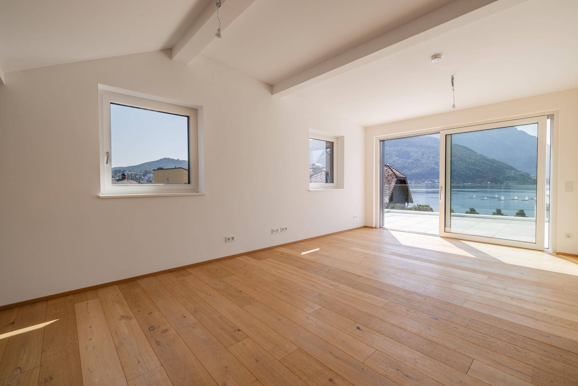 Heller Wohnraum mit Holzboden, großen Fenstern und direktem Blick auf den See und die umliegende Berglandschaft.