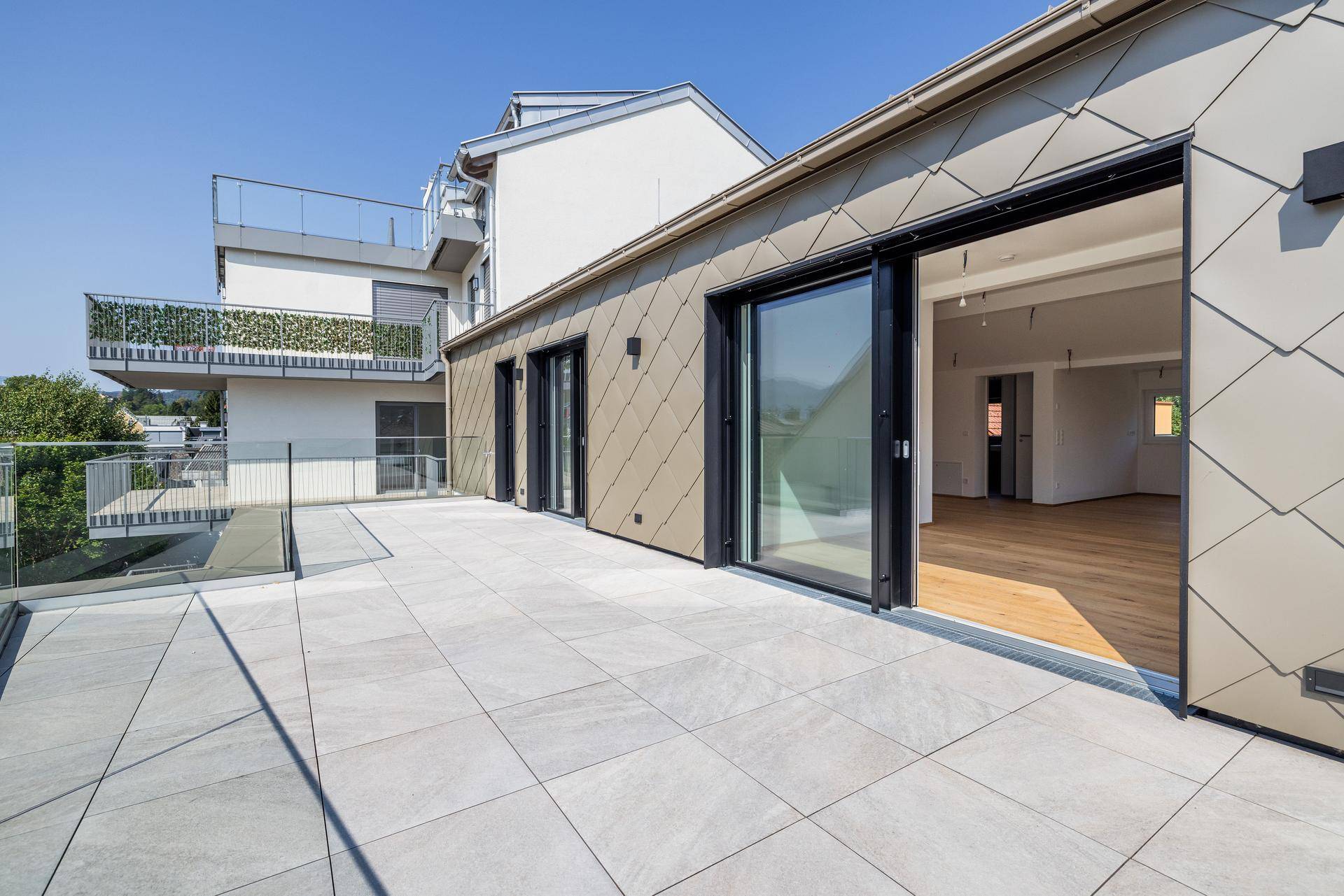 Moderne Terrasse mit Zugang zu einem hellen Innenraum durch große Schiebetüren und schuppenartiger Fassadenverkleidung.