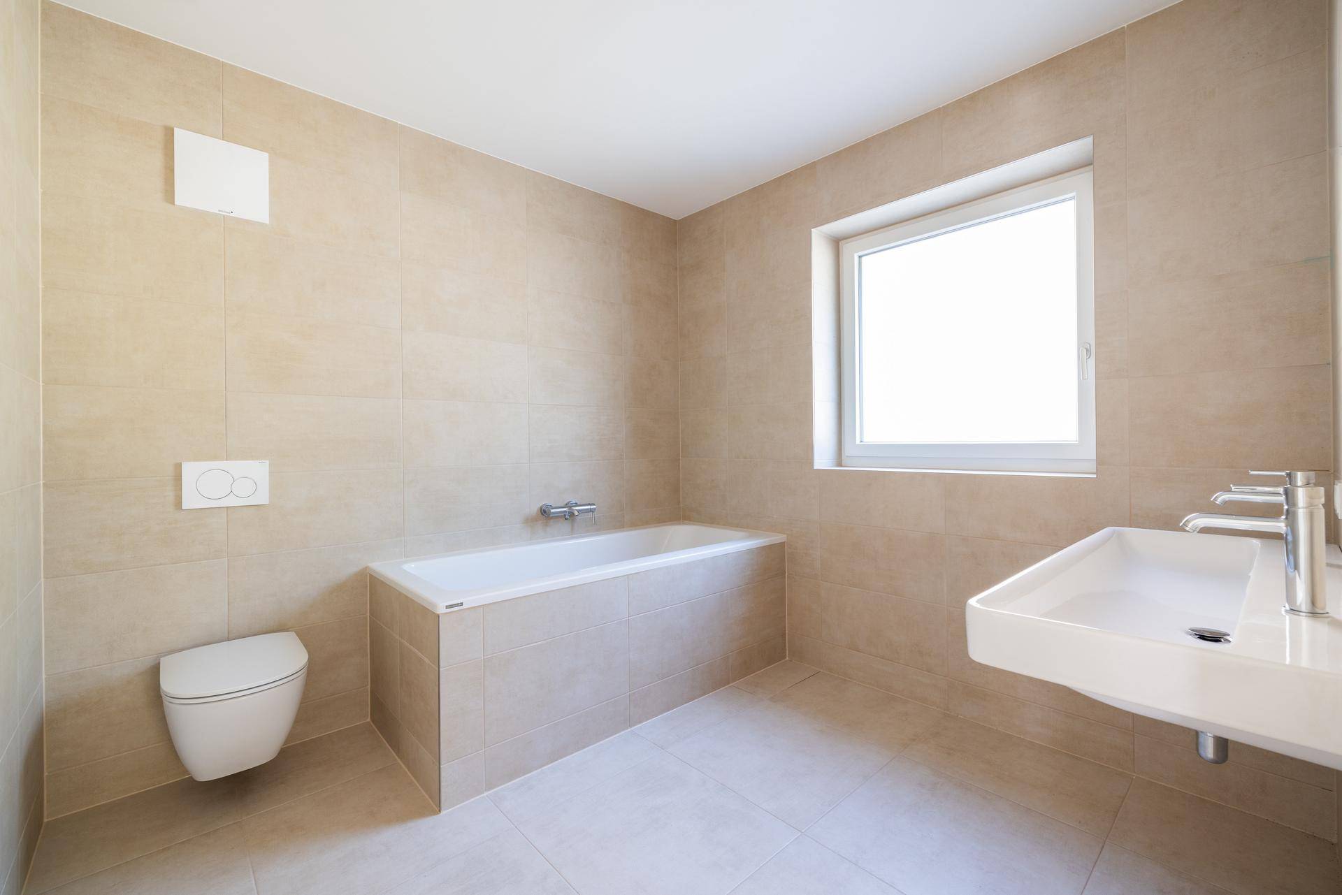 Modernes Badezimmer mit beigefarbenen Fliesen, Badewanne, wandhängendem WC und Fenster für Tageslicht.