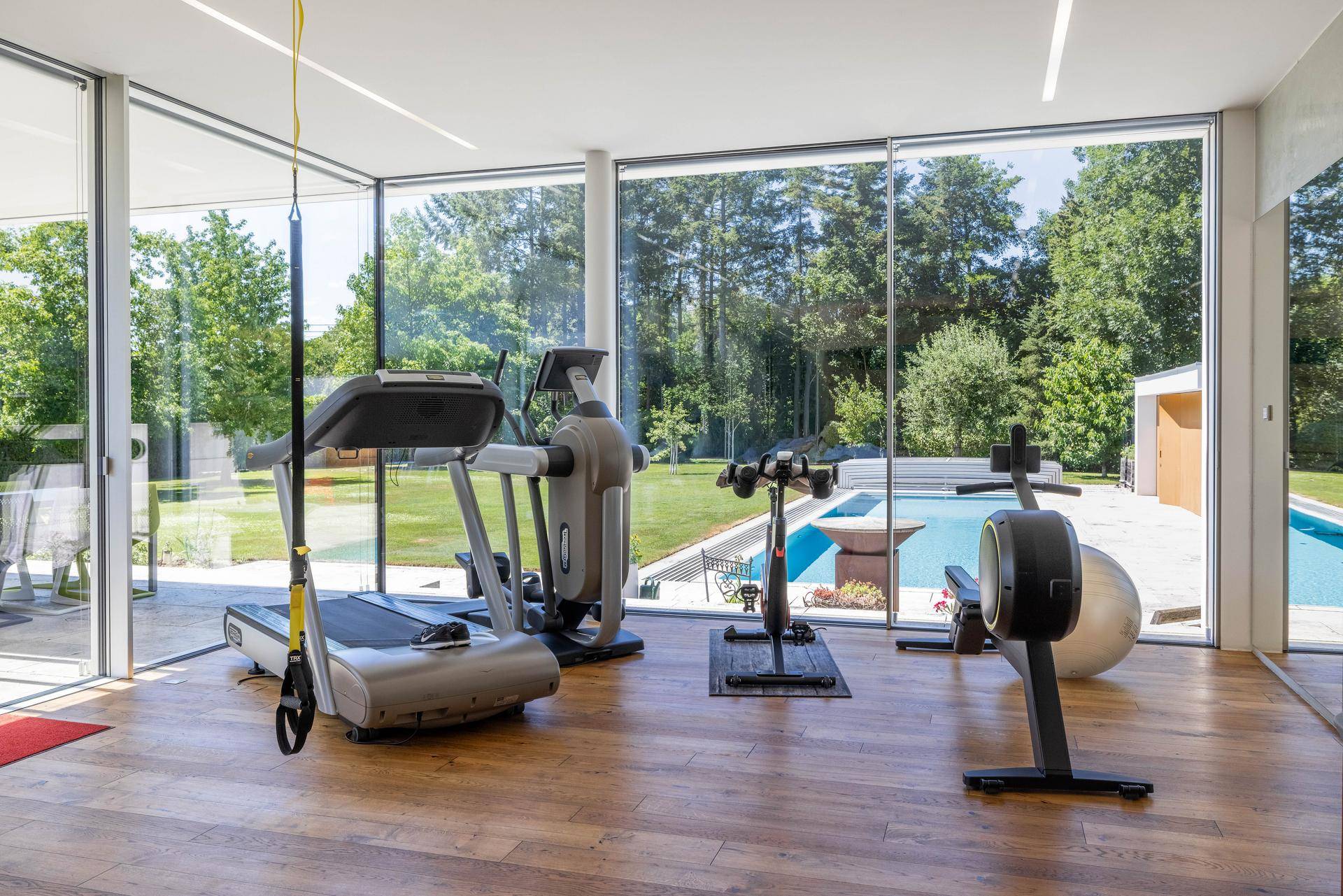 Heller Fitnessraum mit modernen Geräten und Panoramafenstern mit Blick auf den Pool und Garten.