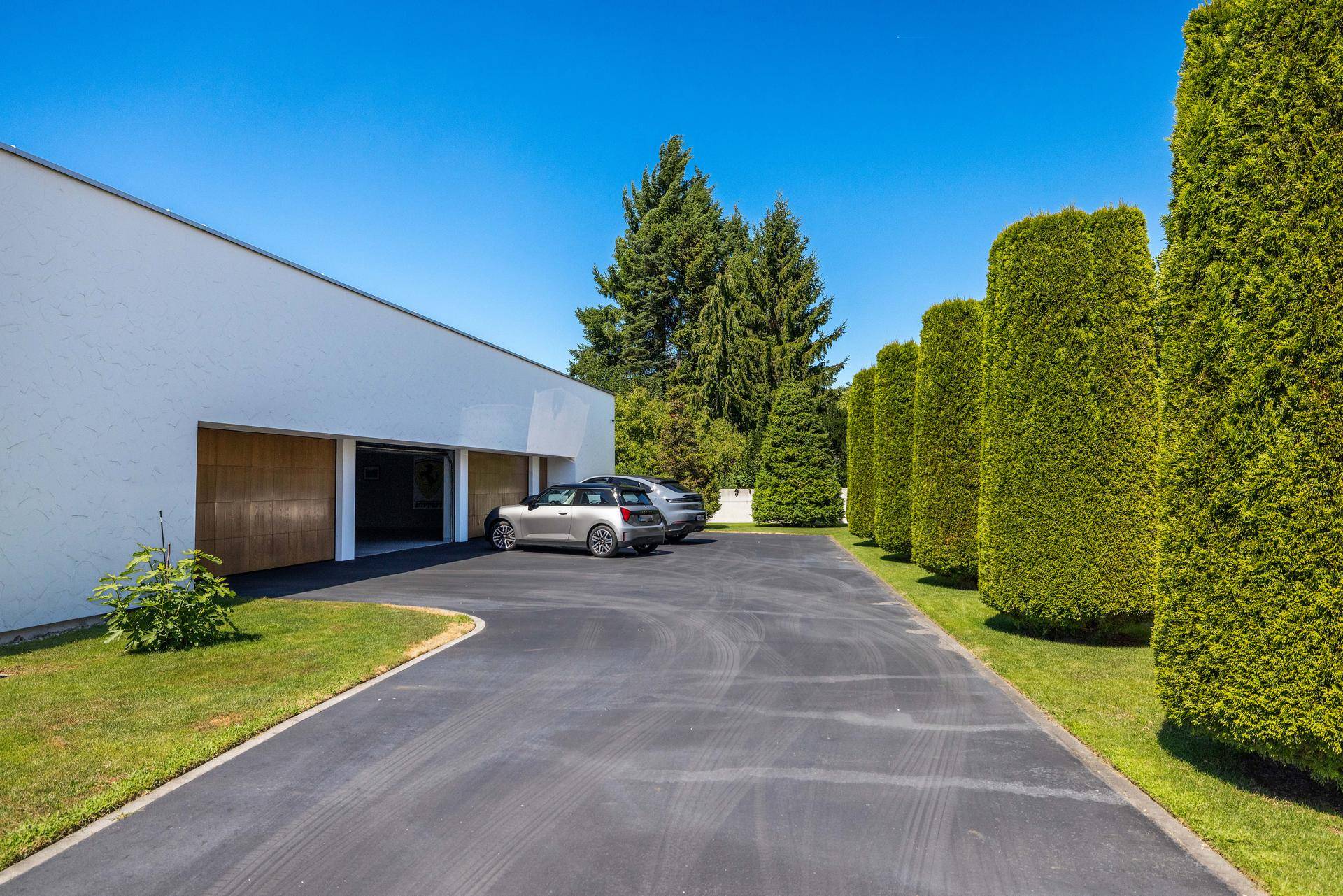 Lange Auffahrt zu einer modernen Garage mit zwei geparkten Autos und hohen Hecken.