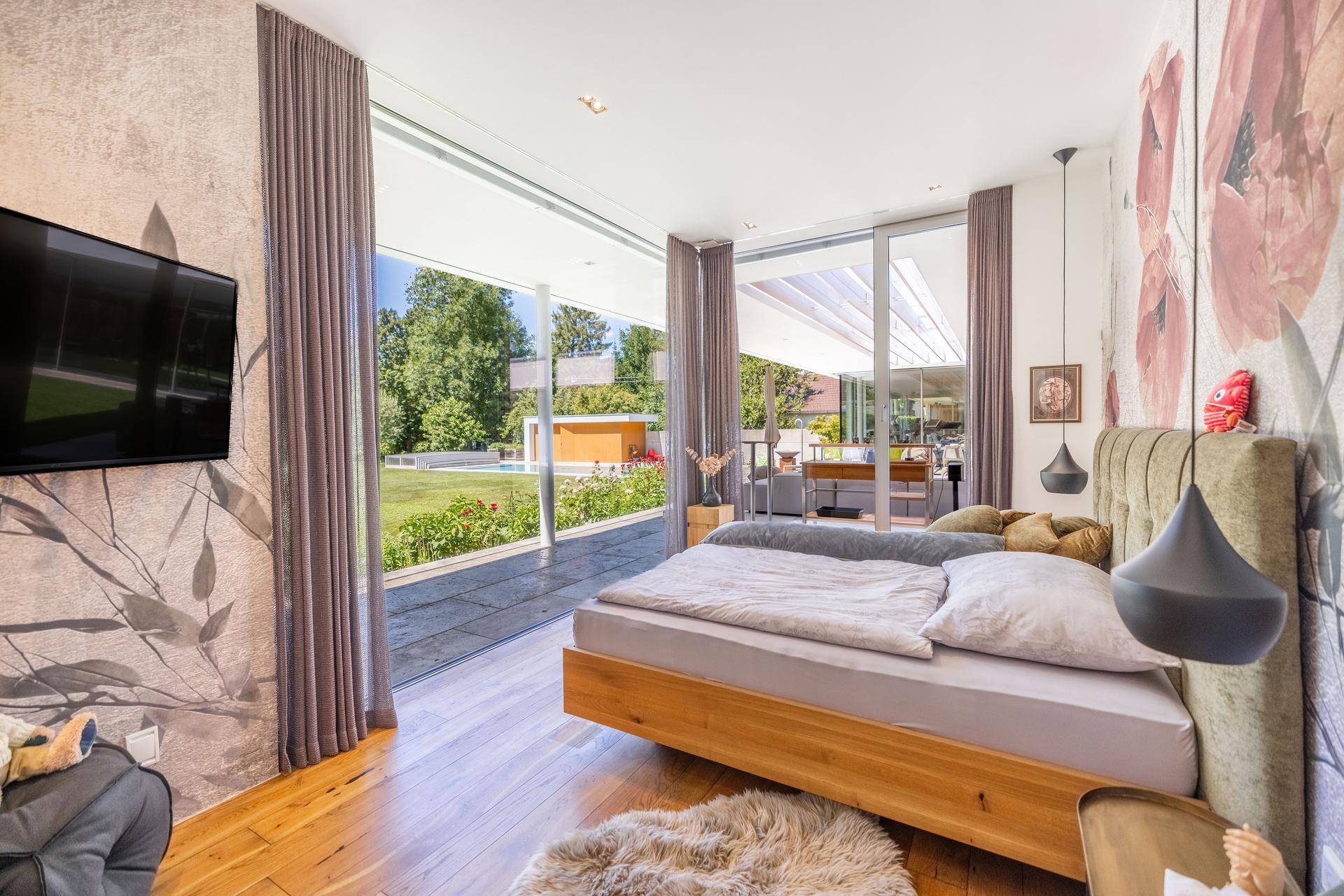 Helles Schlafzimmer mit Holzboden, floraler Tapete und großen Fenstern mit Blick ins Grüne.