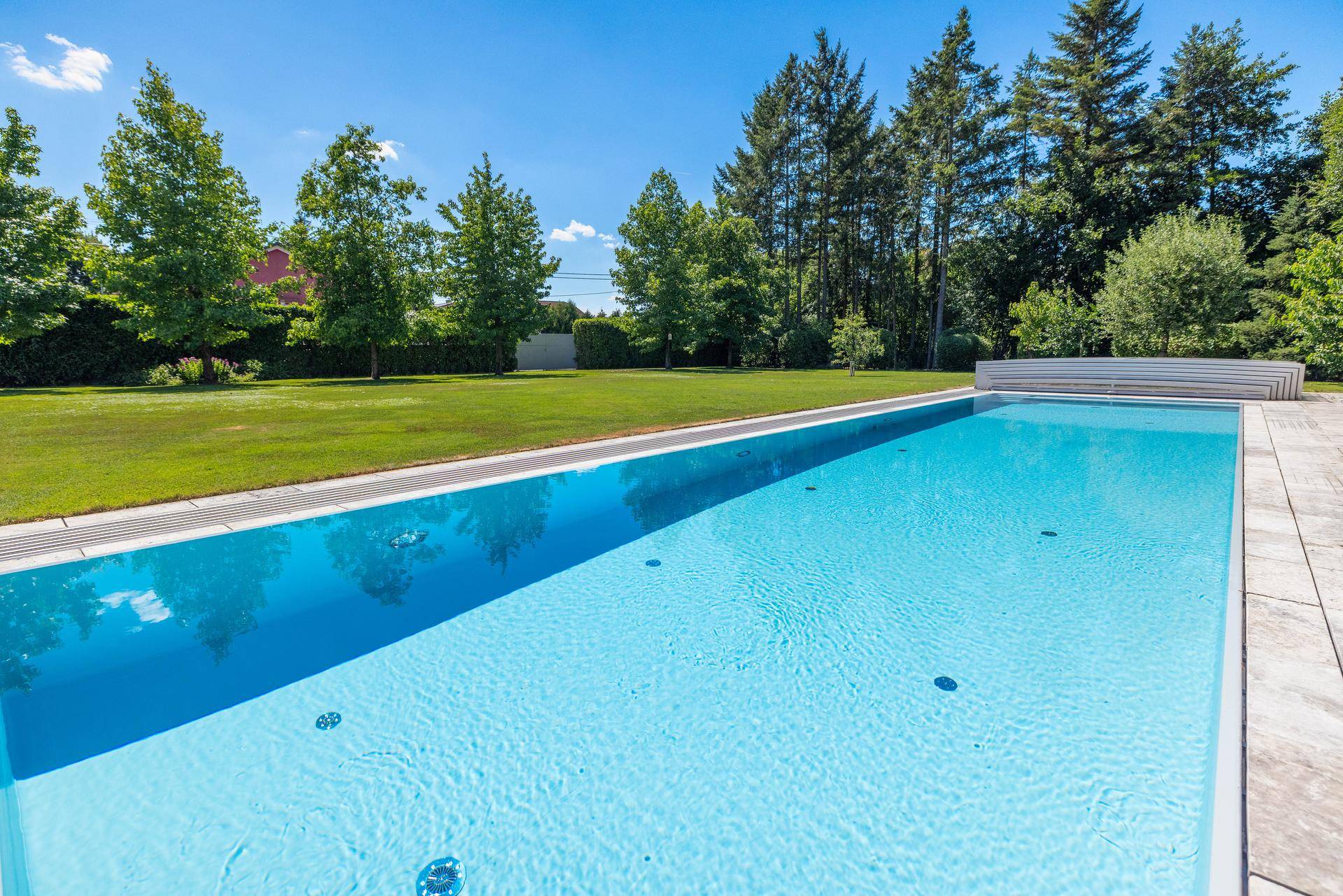 Langer, sauberer Swimmingpool umgeben von einer gepflasterten Fläche und grünem Rasen.