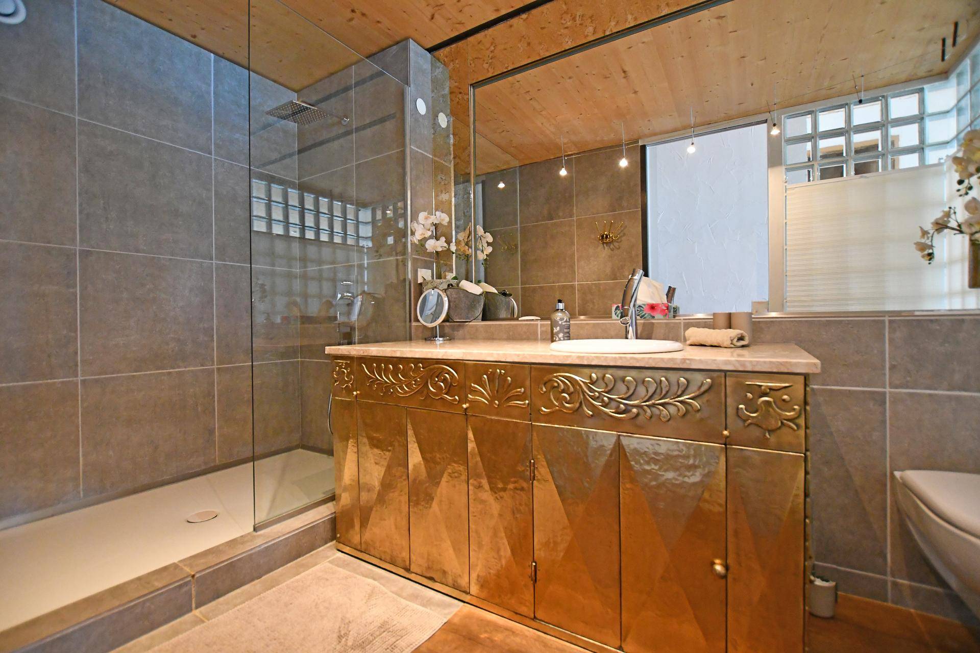Modernes Badezimmer mit begehbarer Dusche, goldenem Waschtisch und Holzbalkendecke.