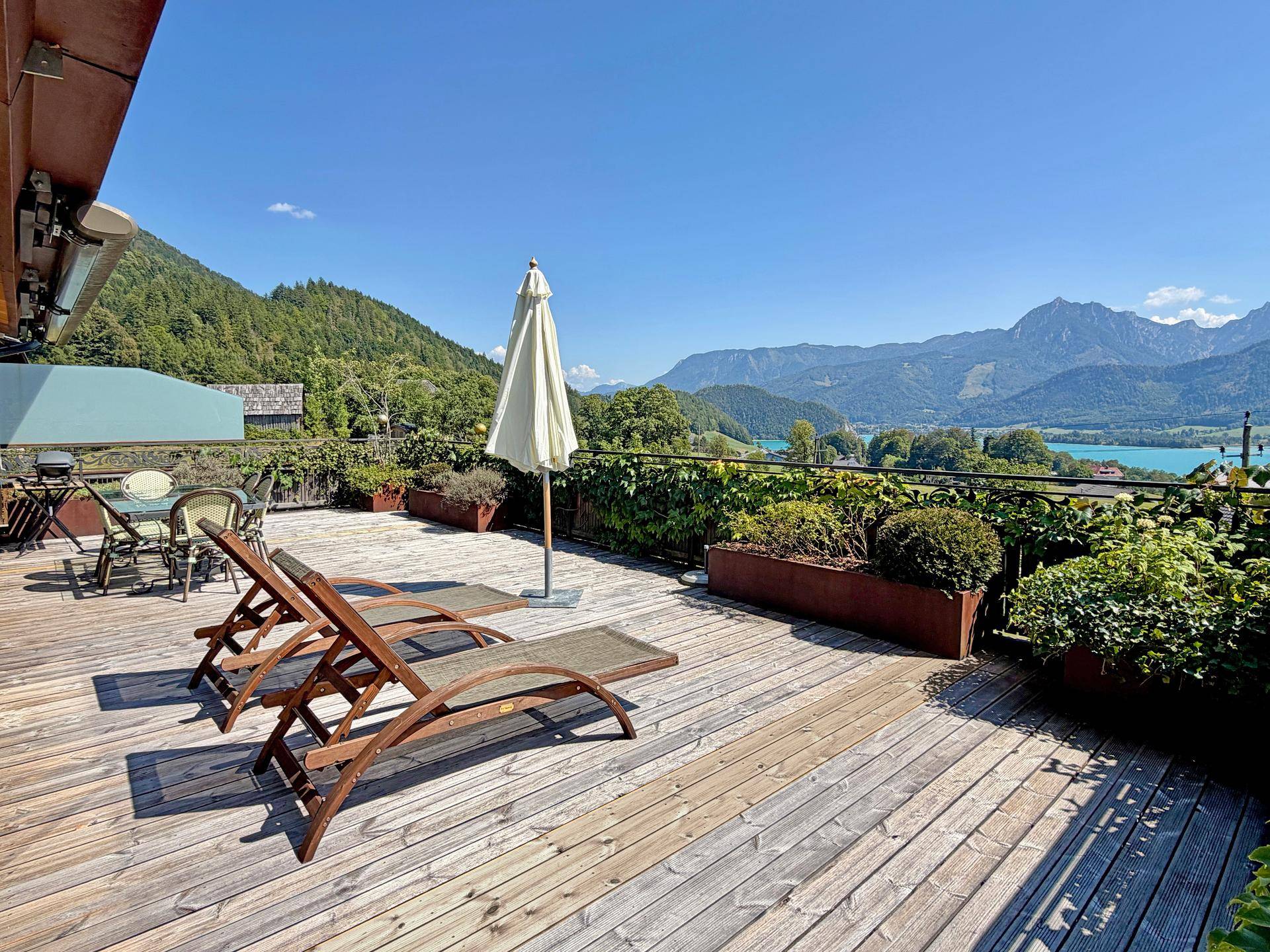 Geräumige Holzterrasse mit Sonnenliegen und Sonnenschirm, bietet einen weiten Blick auf See und Berge.