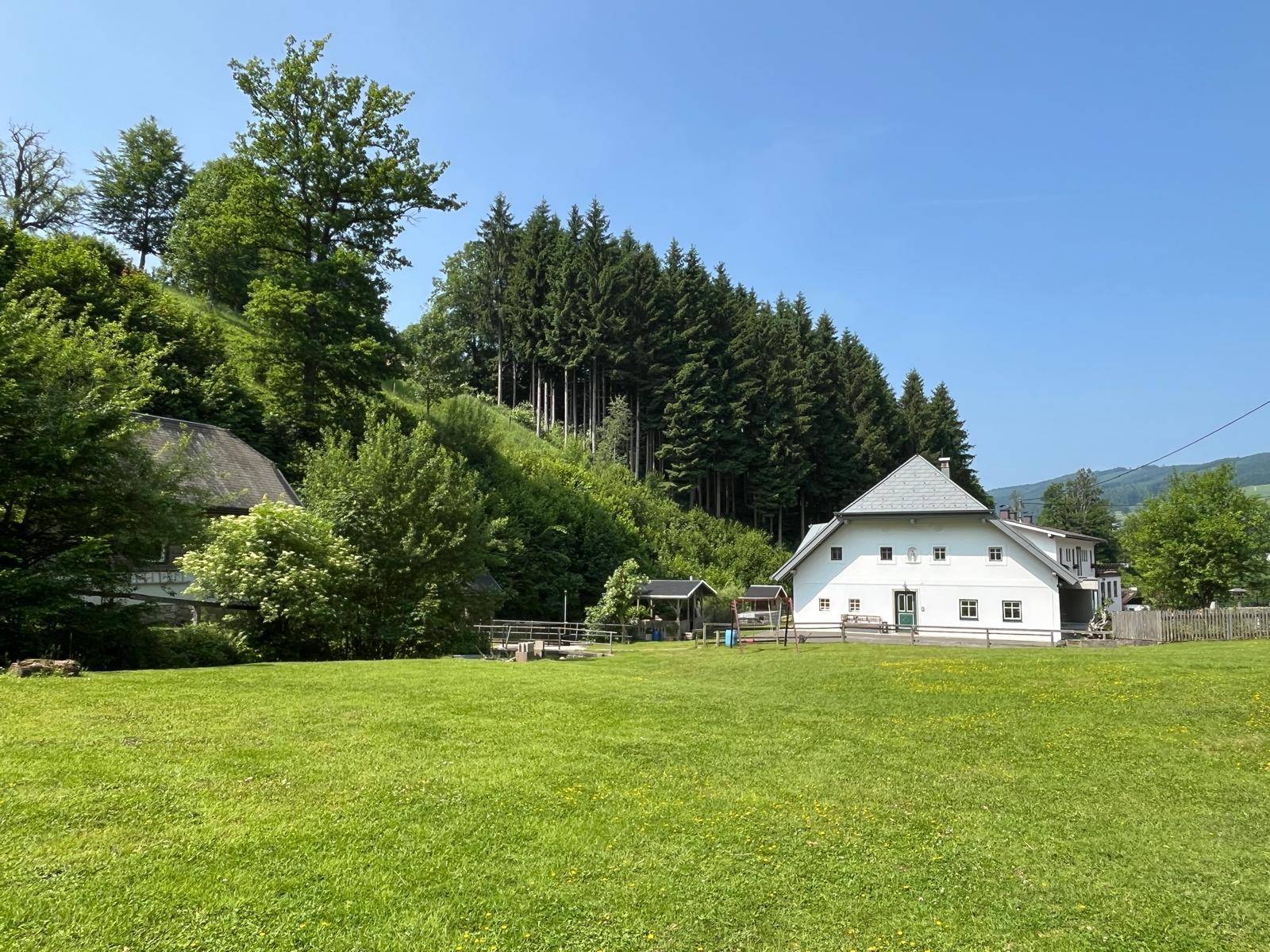 Weitläufige Grünfläche vor einem traditionellen Landhaus mit Wald im Hintergrund.